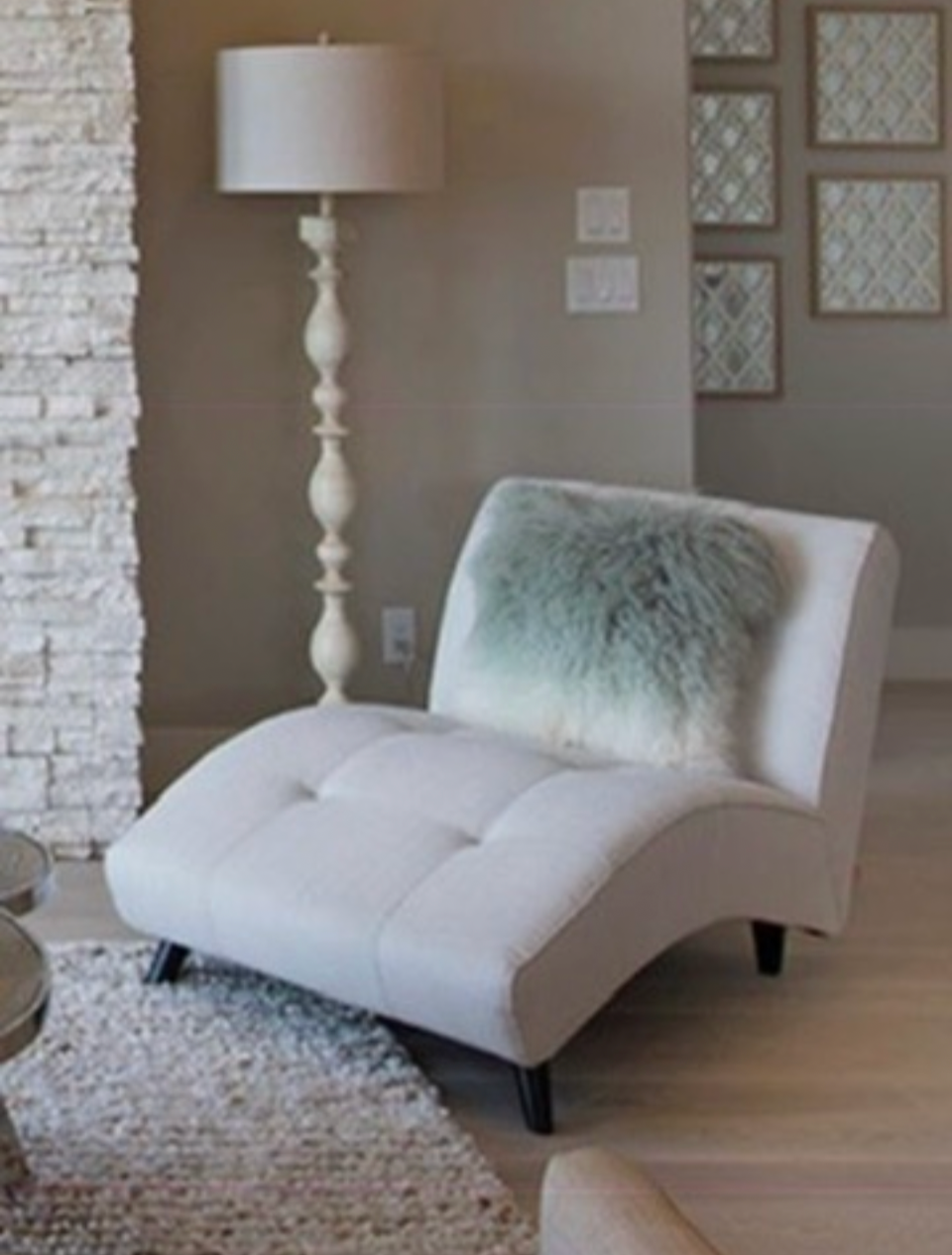 A modern, cream-colored chaise lounge with a furry pillow on it, next to a tall white floor lamp with a beige lampshade, against a wall with framed decorative mirrors. Part of a textured white brick wall is visible on the left.