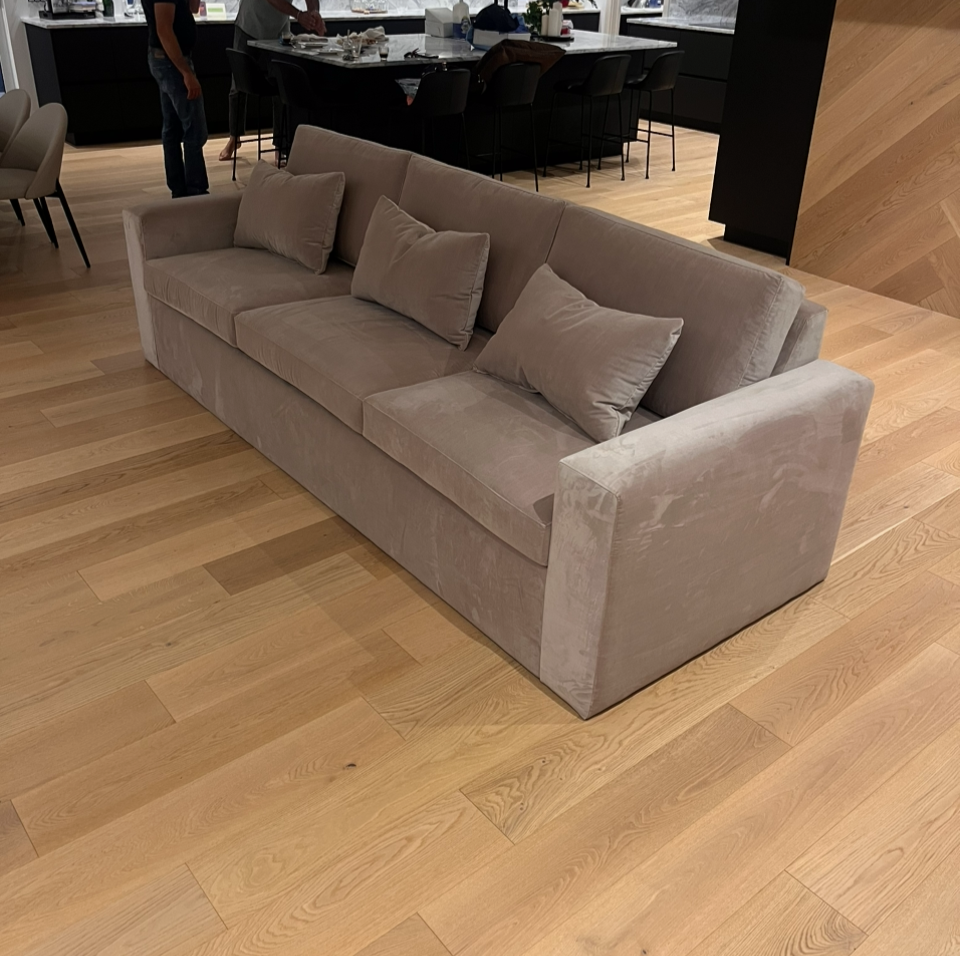 Light beige sofa with three matching cushions in a modern room with wood flooring, alongside a dining area and people in the background.