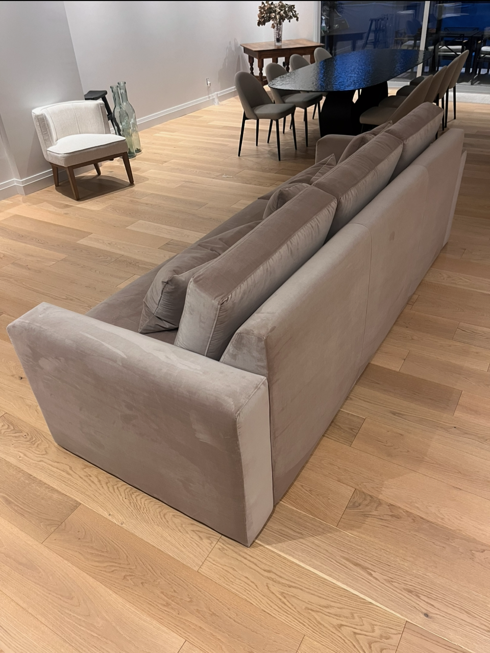 Light beige sofa in a modern living room with hardwood floors, near a dining area with a dark table and gray chairs, and a small white armchair with wooden legs and glass bottles beside it.