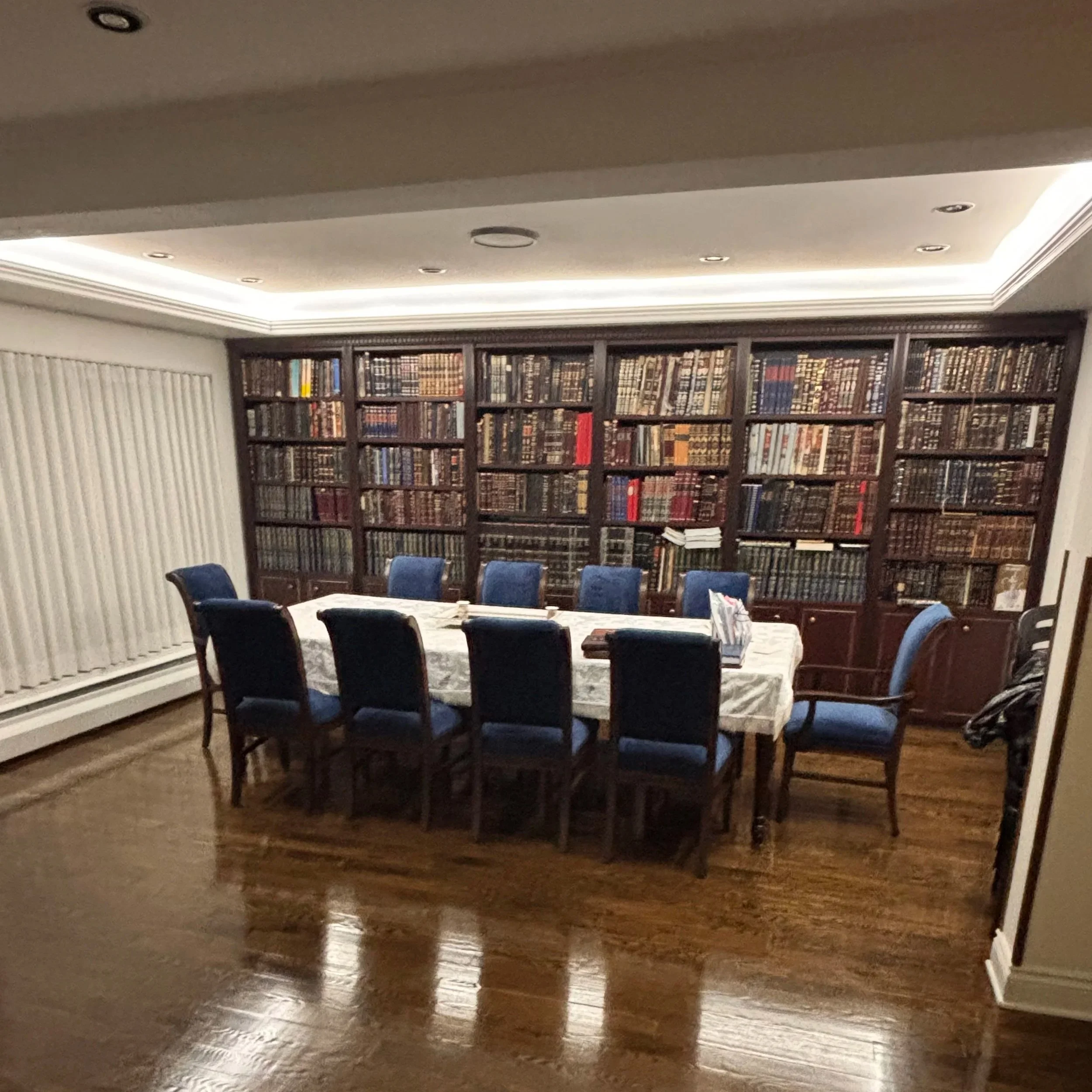 A dining room with a large wooden table covered with a white tablecloth, surrounded by ten blue upholstered chairs, and a large dark wood bookcase filled with books against the wall.