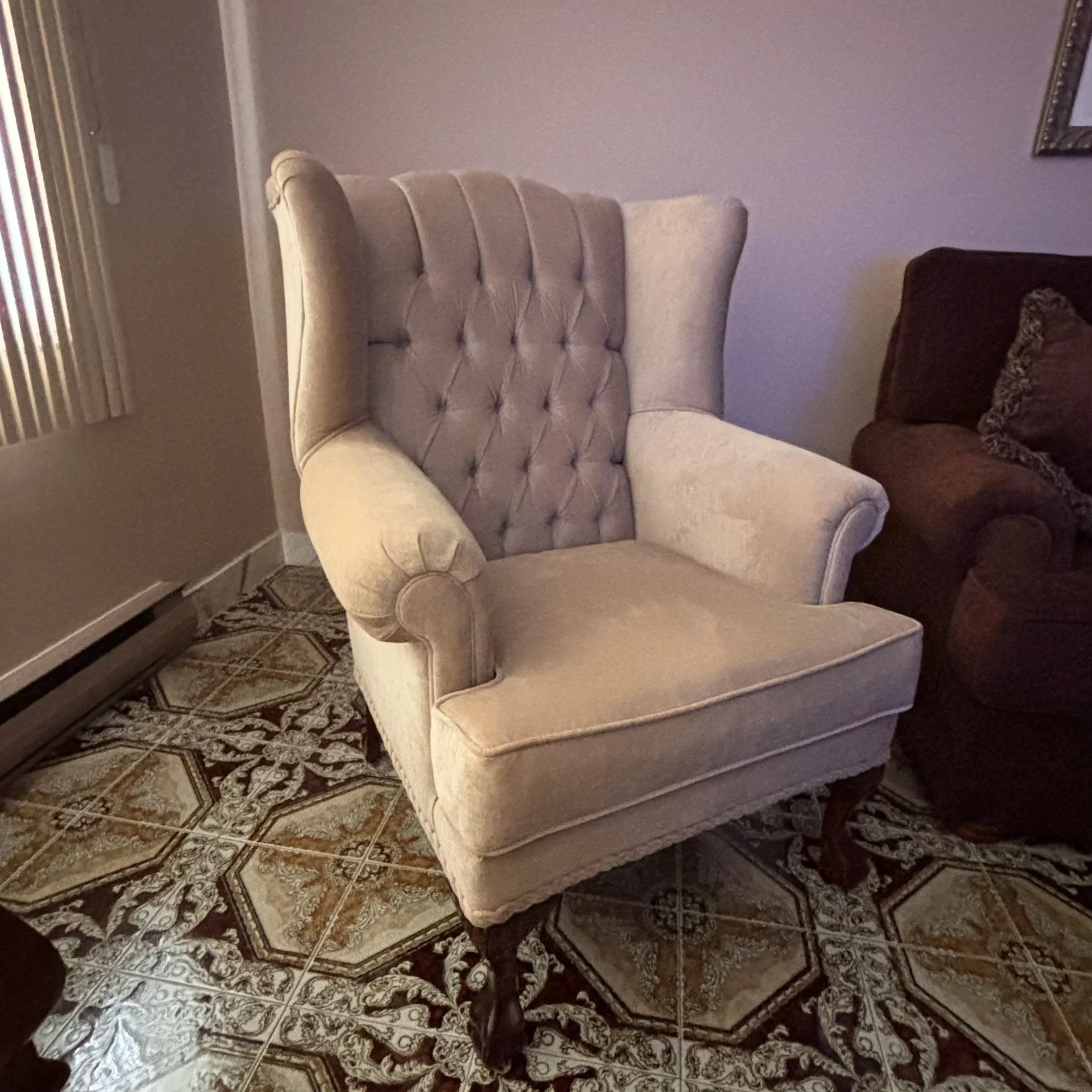 An elegant beige wingback armchair with tufted backrest and wooden legs, placed on a patterned tile floor in a living room.
