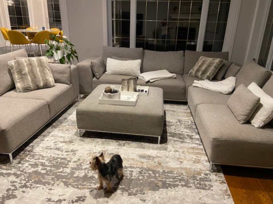 Living room with beige sofas, throw pillows, a large gray ottoman, and a small dog walking on a patterned rug; in the background, a dining area with yellow chairs and a table.