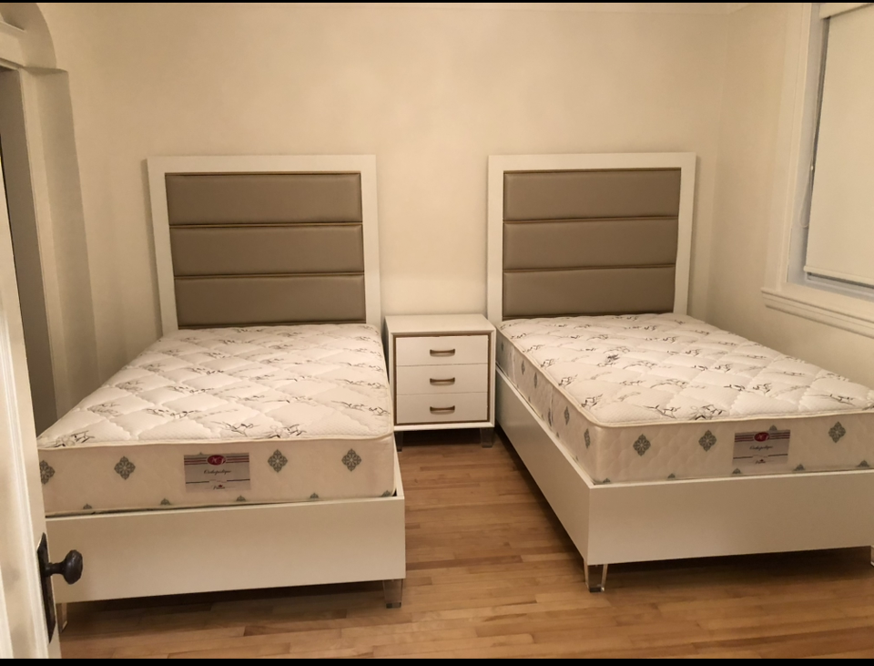 Two twin beds with padded headboards and a white nightstand between them in a room with hardwood floors and a window with closed blinds.