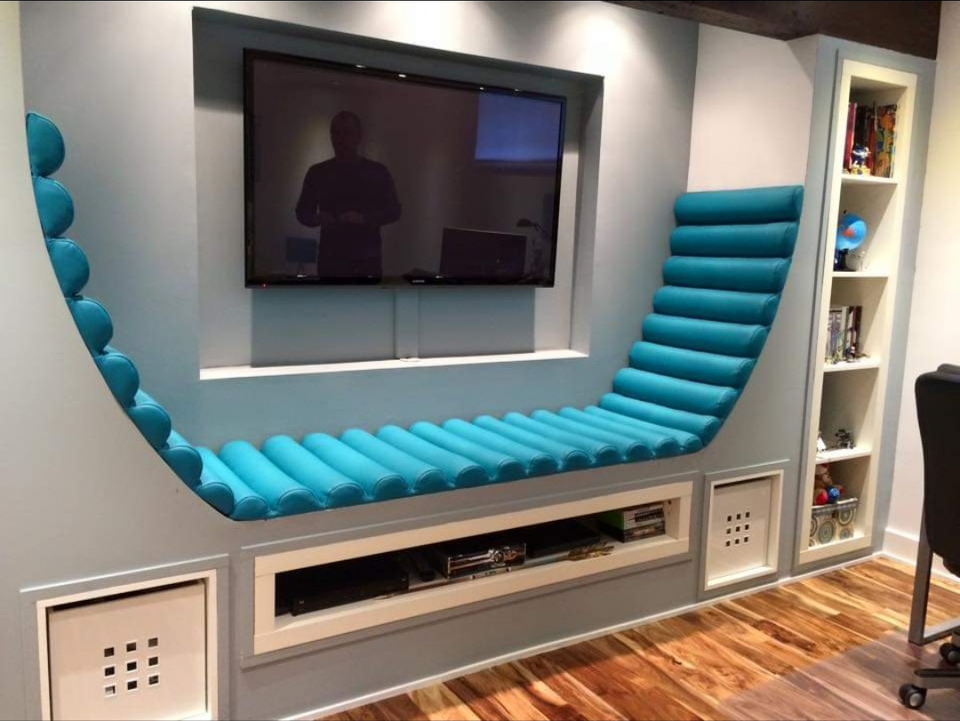 A built-in wall entertainment center with a mounted flat-screen TV in a modern living room. The design features a curved, padded, blue seat around the TV nook, with white shelving and storage units, and hardwood flooring.