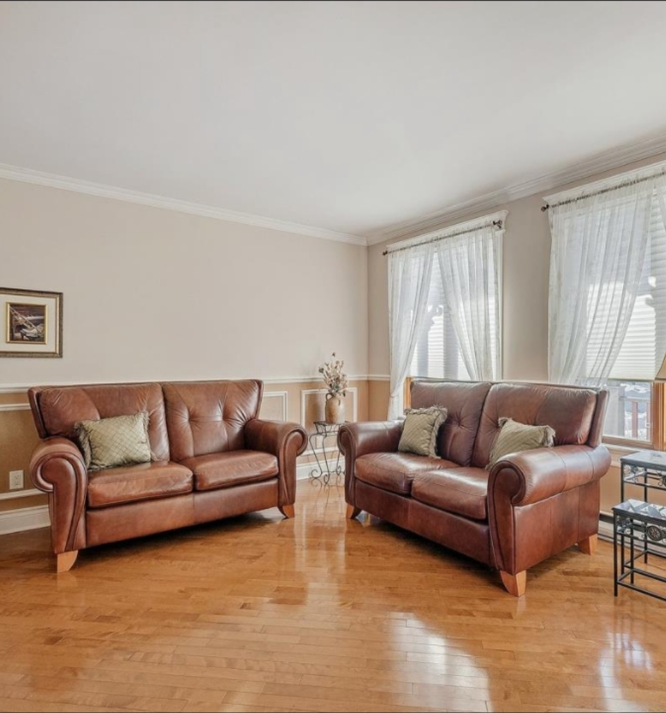 Living room with two brown leather sofas, a side table with a vase and flowers, and large windows with white curtains.