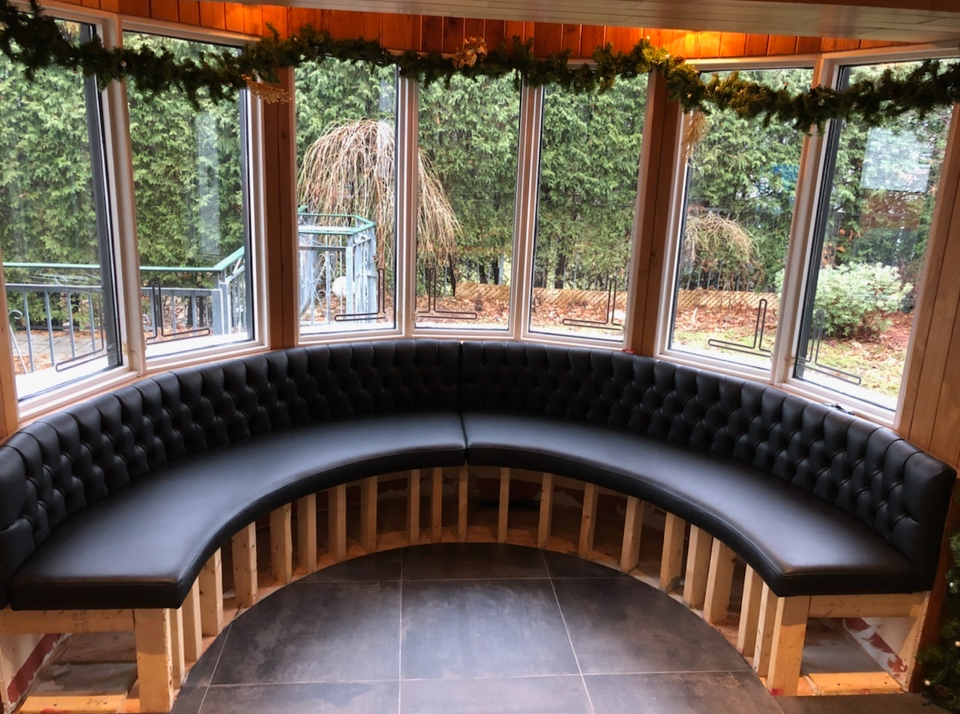 A cozy, semi-circular black leather cushioned bench inside a wooden room, with large windows showing a backyard with trees, bushes, and a patio. Christmas garland decorates the top of the windows.