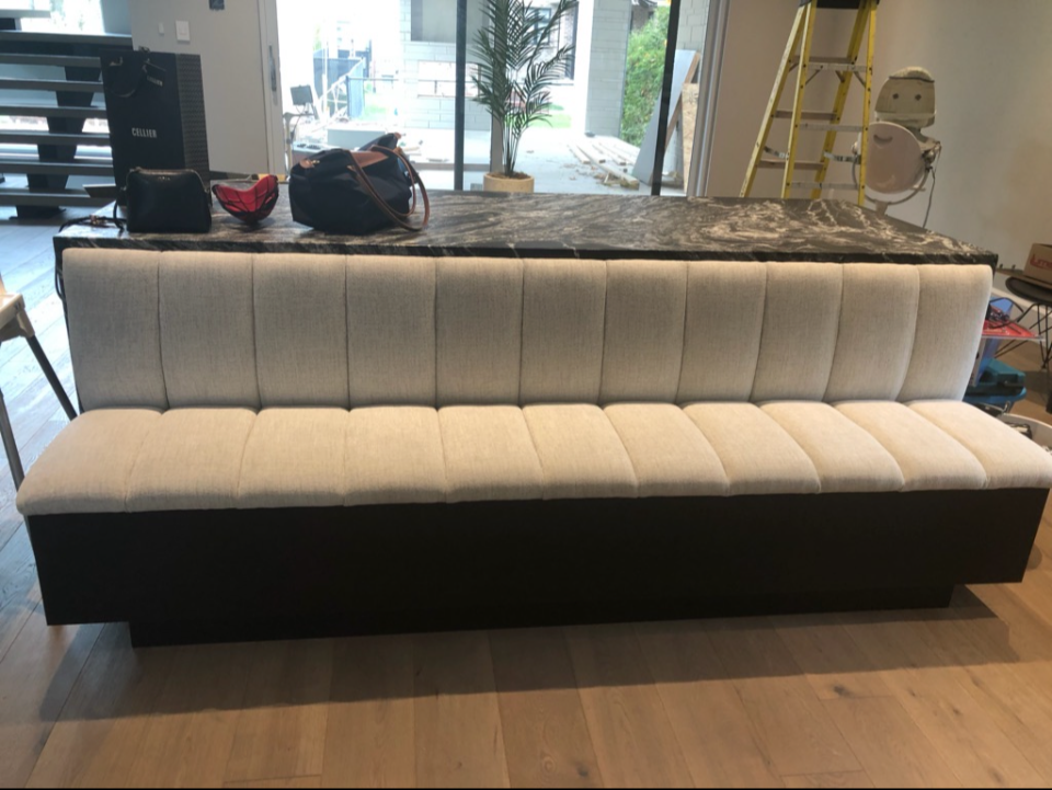 A beige upholstered sofa with vertical stitching is positioned in front of a black marble countertop in a modern interior space. Items including handbags and personal belongings are placed on the counter. Behind the sofa, a large glass door or window