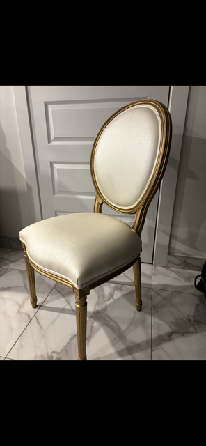 A cream-colored upholstered chair with a rounded back and gold trim, placed on a tiled floor in front of a white door.