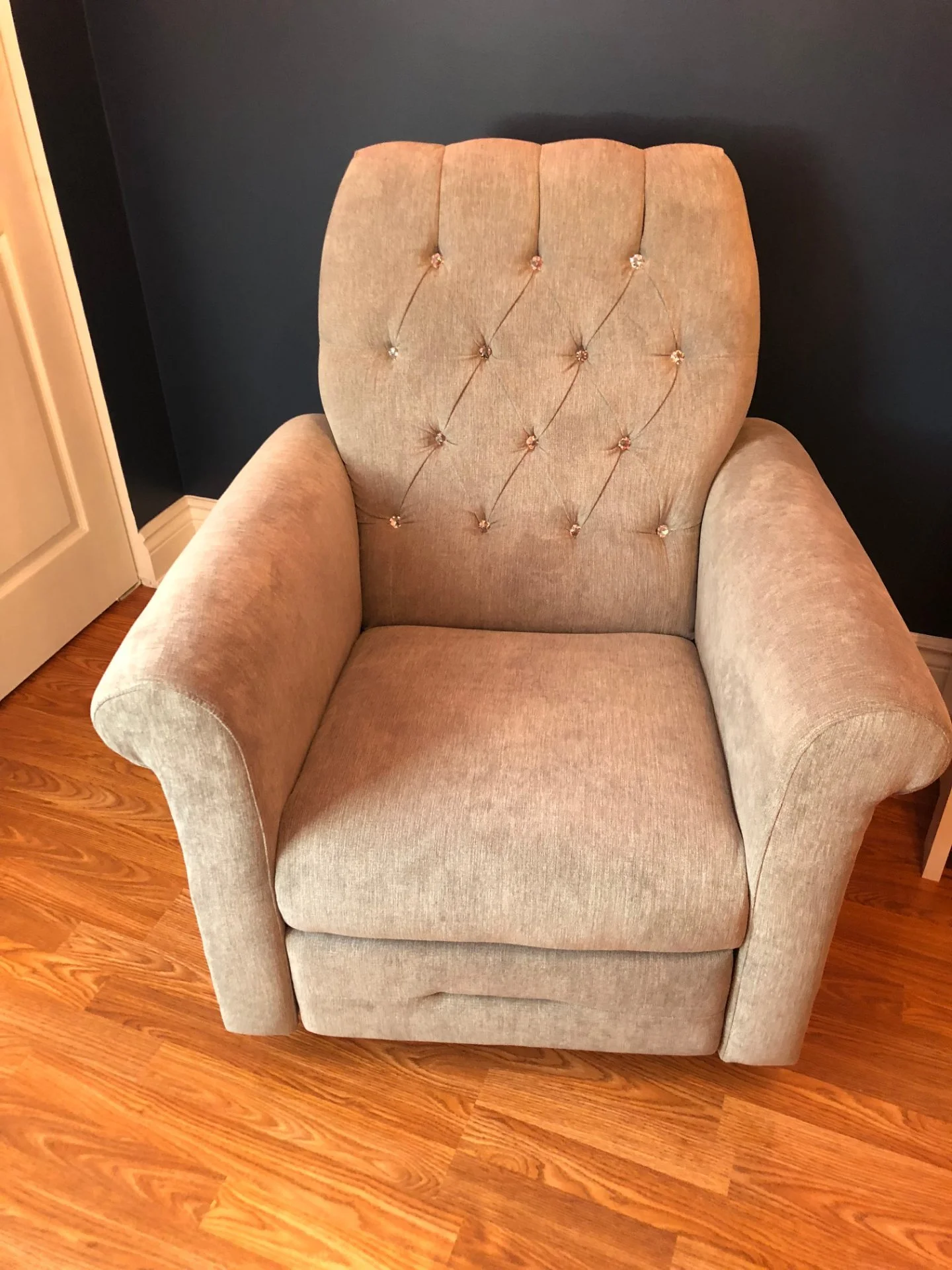Beige upholstered armchair with tufted backrest and crystal button accents, placed on hardwood floor against a dark wall.