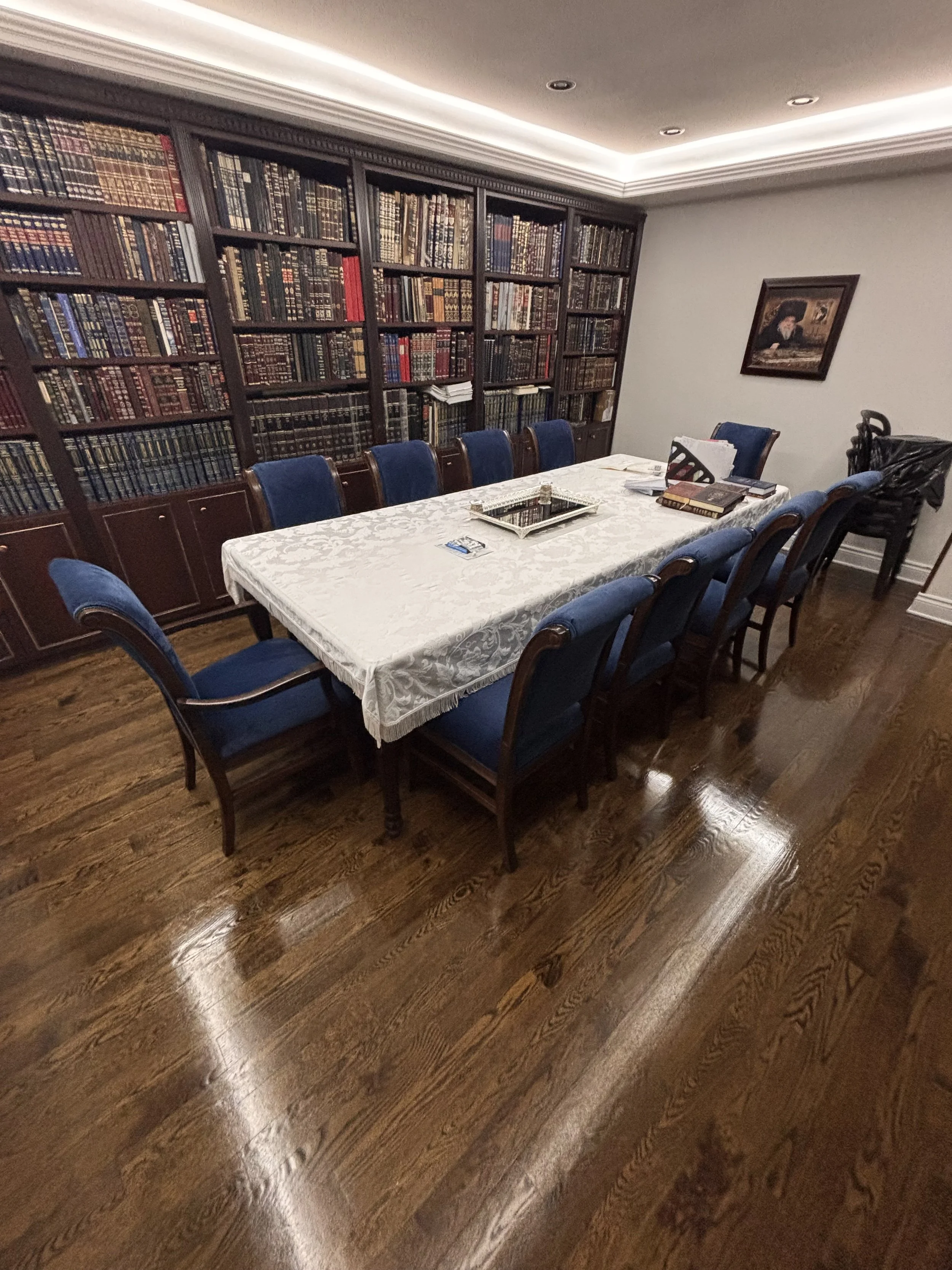 A dining room with a large wooden table covered by a white tablecloth. Eight blue-upholstered chairs are around the table. A large bookshelf filled with leather-bound books lines the back wall, and a framed painting hangs on the side wall. The room h