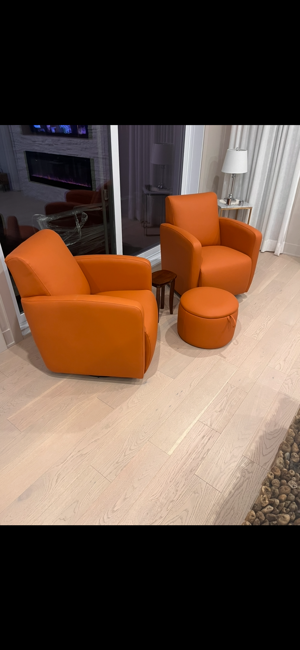 Two orange leather armchairs and a matching ottoman in a living room, with a small wooden side table between them, a window with curtains behind, and a fireplace with a television above it.