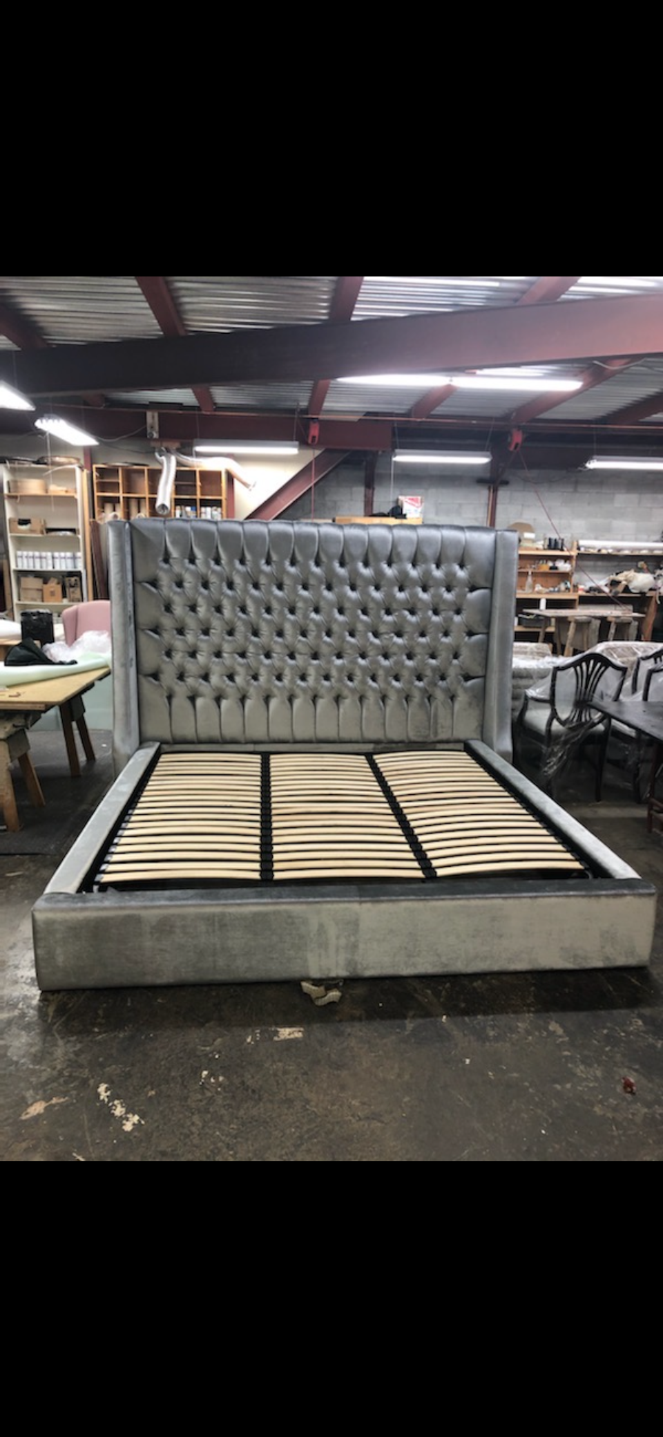 A grey upholstered bed frame with a tufted headboard in a furniture workshop.