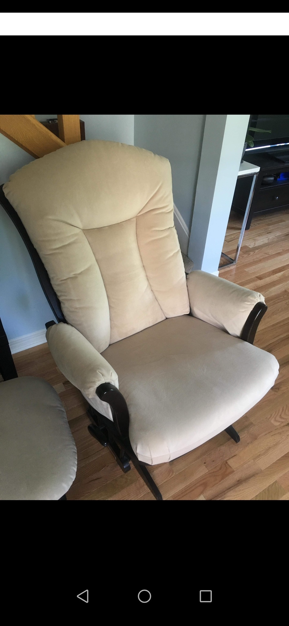 A beige upholstered armchair with dark wooden armrests and a high back, positioned on a wooden floor next to a small beige cushioned stool.