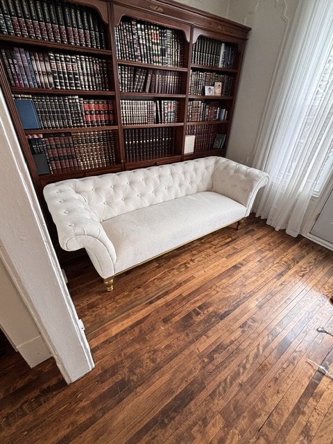 A white velvet tufted sofa with gold feet in front of a large wooden bookcase filled with books, next to a window with white curtains and wood flooring.