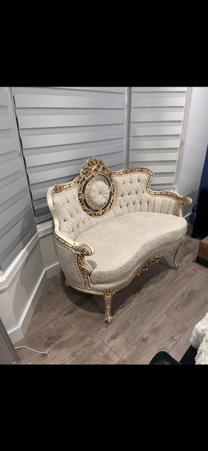 Vintage cream-colored tufted loveseat with gold carved wood accents and a small circular mirror on the backrest, placed against a white panel wall in a room with hardwood flooring.
