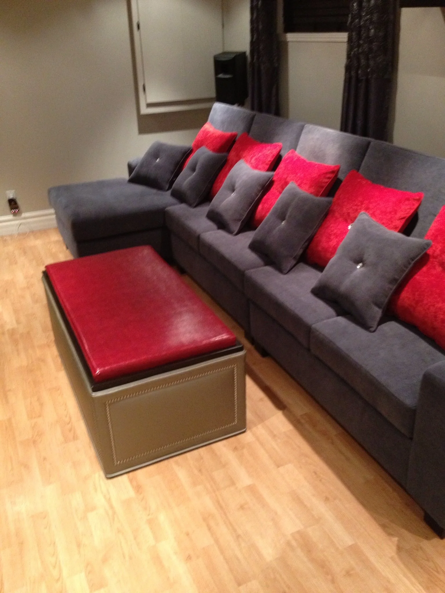 A living room with a gray sectional sofa, red and gray throw pillows, a red leather ottoman, wooden flooring, a window with dark curtains, and a wall-mounted speaker.