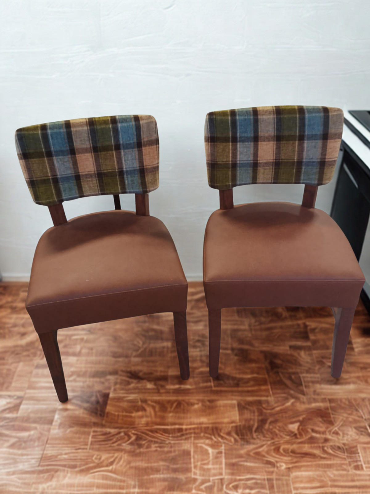 Two vintage chairs with plaid fabric backs and brown upholstered seats, positioned on a wooden floor against a white wall.