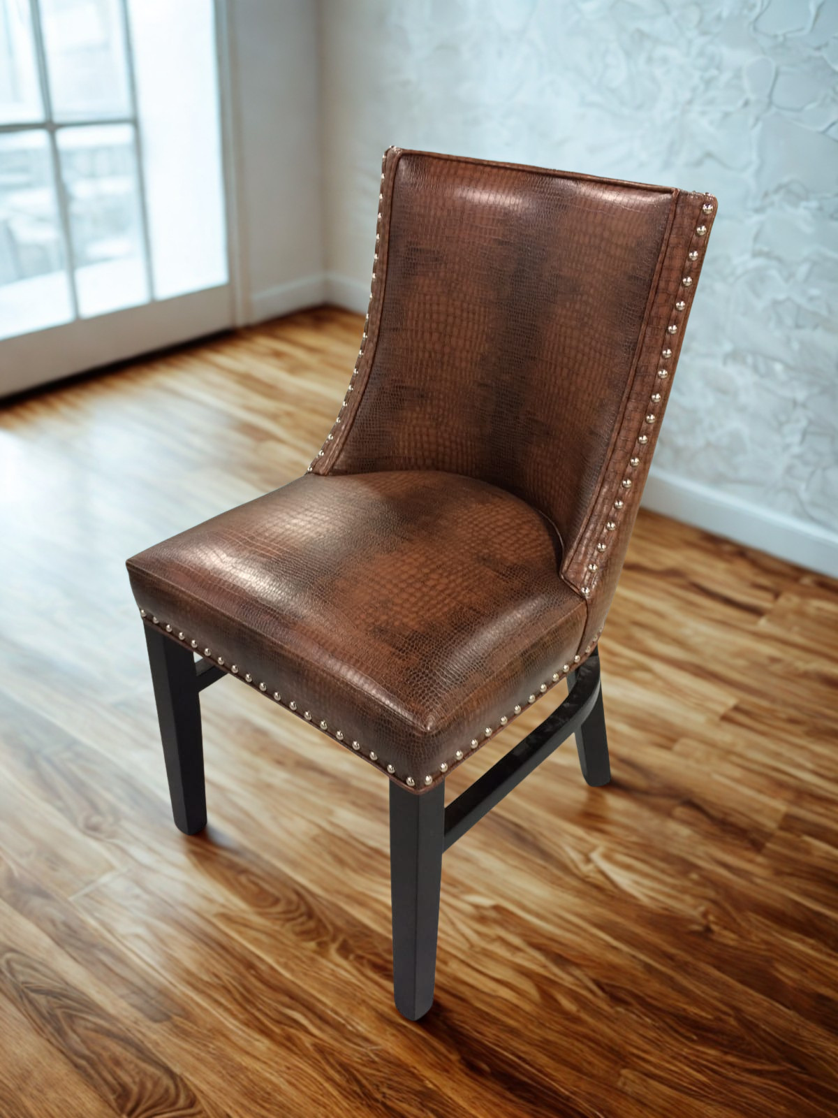 Brown leather upholstered chair with nailhead trim on a wooden floor near a window.