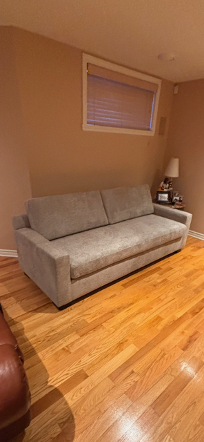 A gray upholstered sofa with a backrest and armrests in a living room, next to a window with a closed blind and a side table with a lamp.