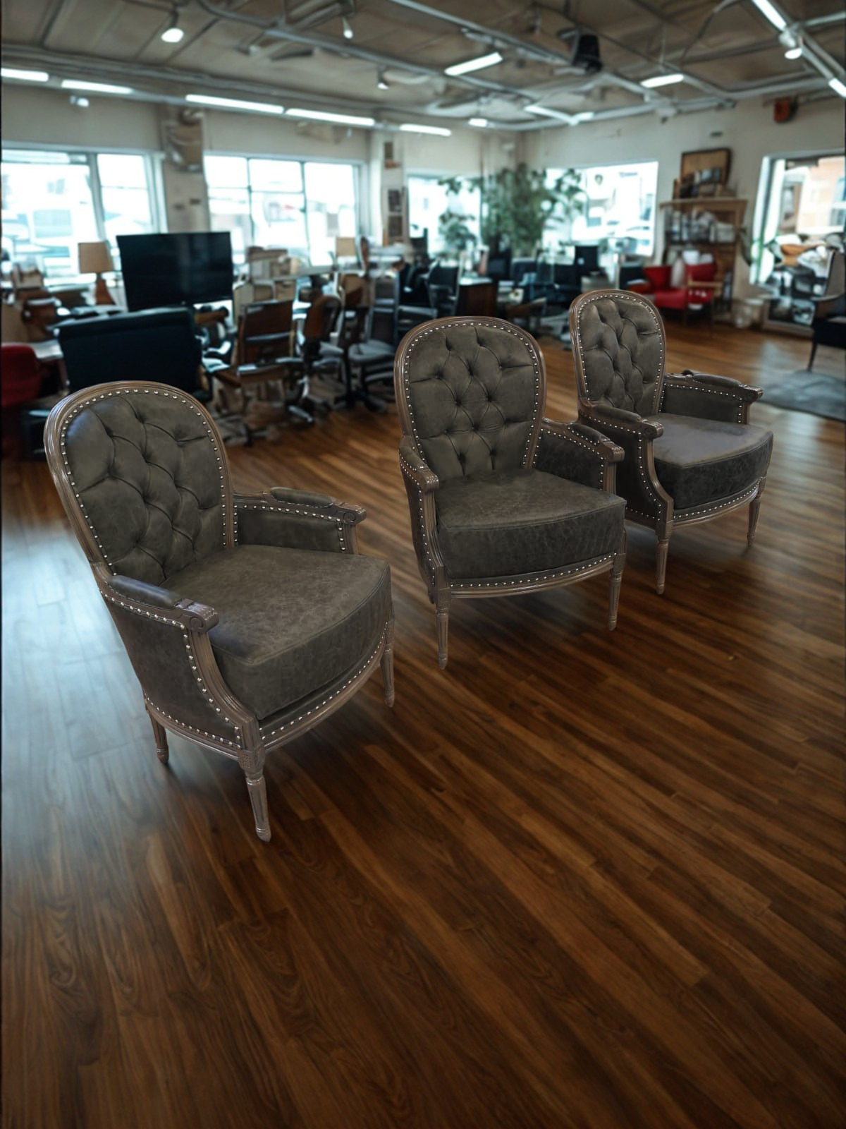 Three vintage armchairs with tufted backs and wooden frames in the foreground of a furniture store with wooden floors and various chairs, tables, and decor in the background.