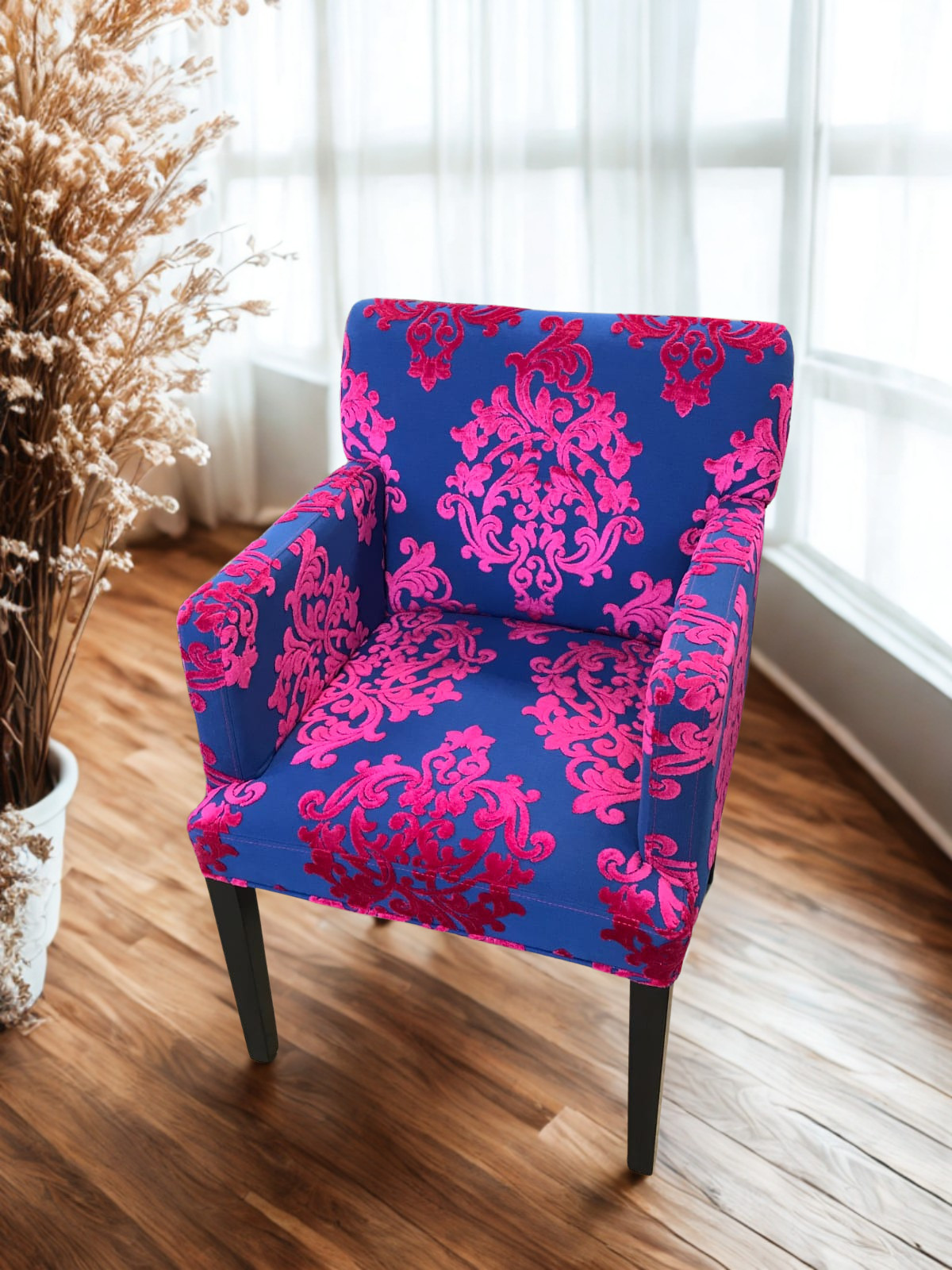 A blue upholstered armchair with pink floral patterns, positioned on a wooden floor near a large window with sheer curtains, with dried plants in a white pot nearby.