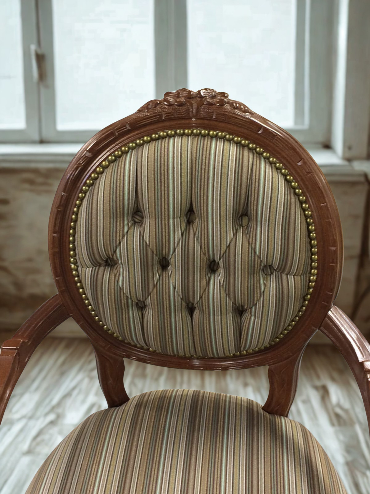 A vintage upholstered wooden chair with a striped fabric pattern and gold nailhead trim, positioned in front of a window.