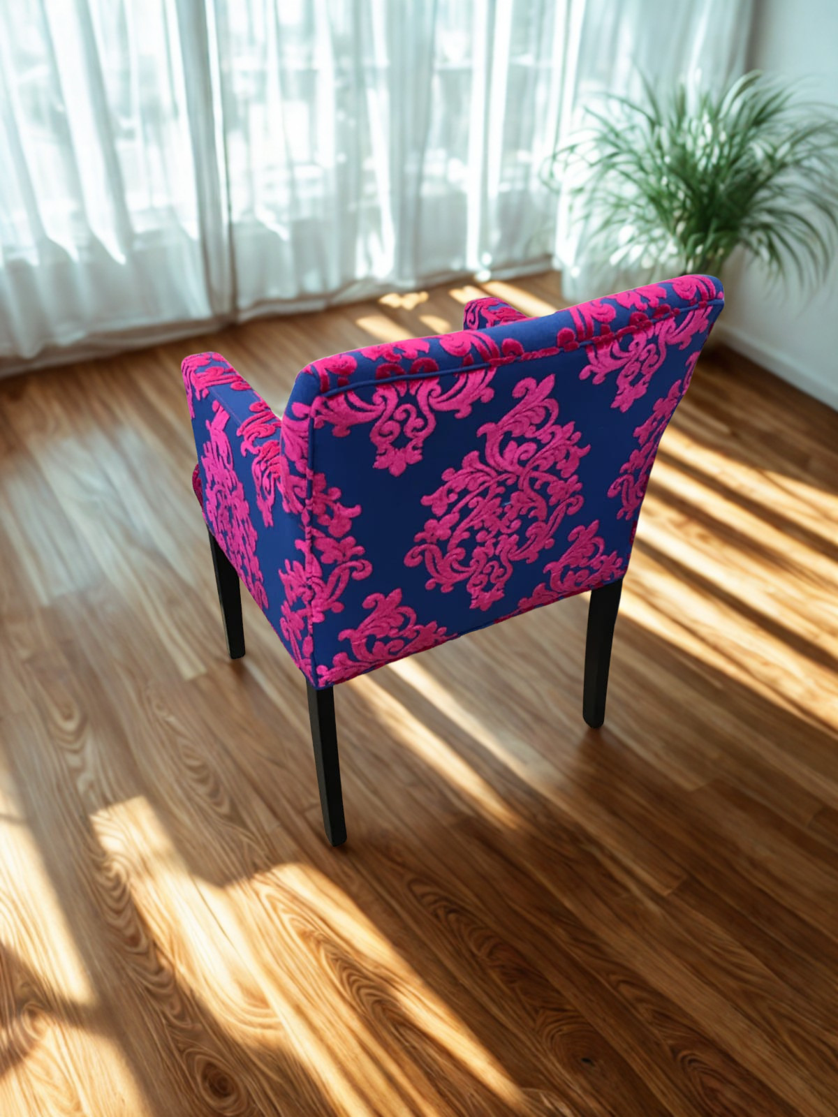 A blue armchair with pink floral patterns in a brightly lit room with wooden flooring and sheer white curtains, next to a large green potted plant.
