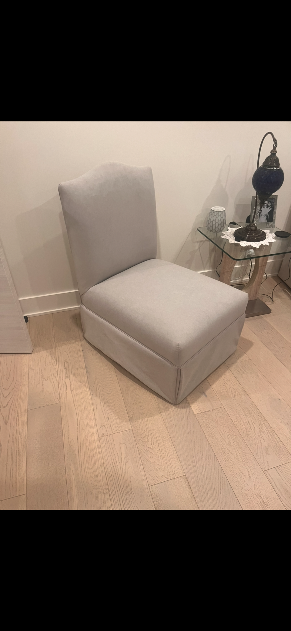A beige upholstered armchair with a high back, positioned next to a glass side table with wooden legs. The table holds a lamp, a small decorative vase, a photograph frame, and an electronic device. The room has light wood flooring and white walls.