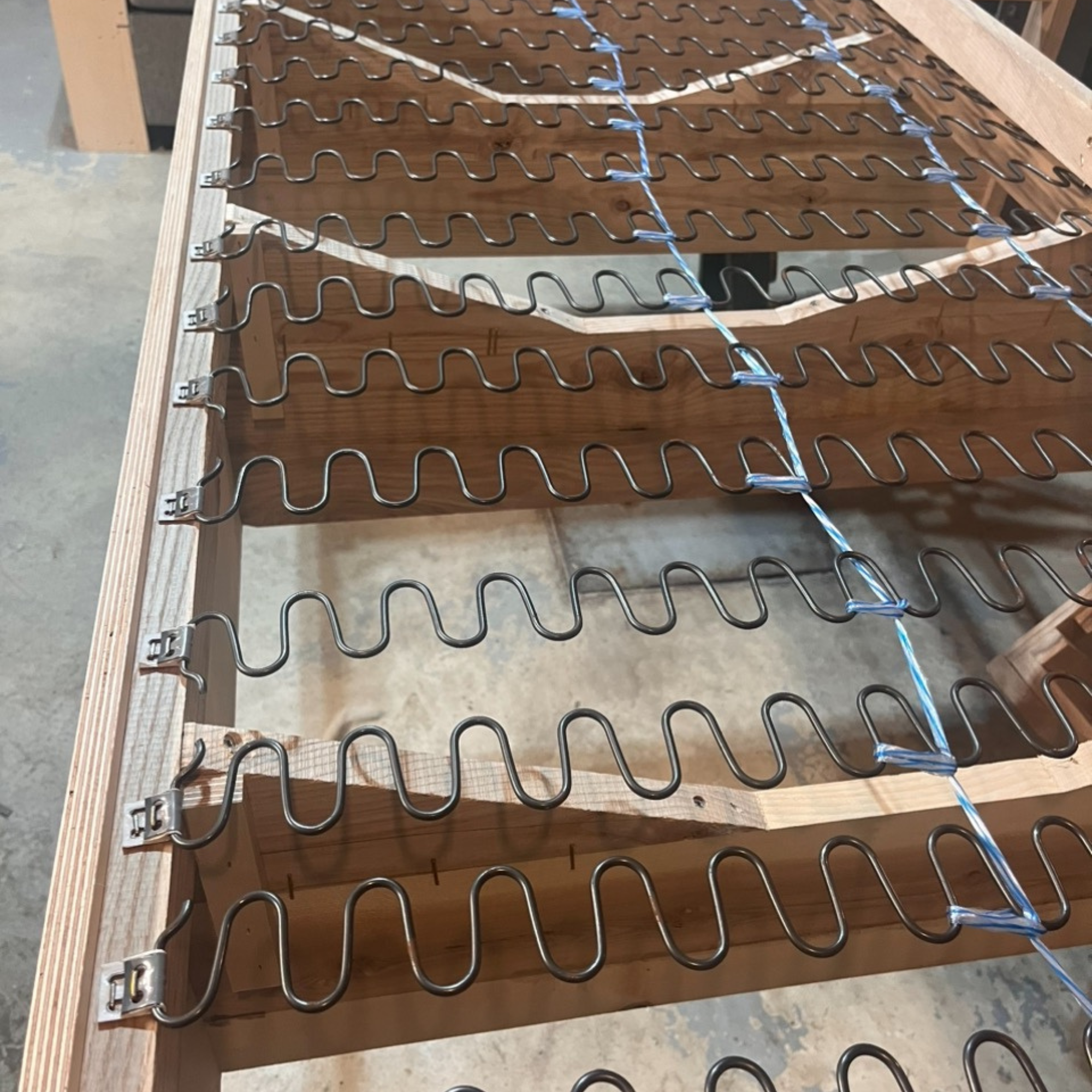A wooden frame with metal coil springs attached, likely part of a mattress framework in a woodworking or furniture shop.