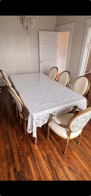 A dining room with a wood floor, six cream-colored chairs with gold trim around a rectangular table covered with a lace tablecloth, and a chandelier hanging from the ceiling.