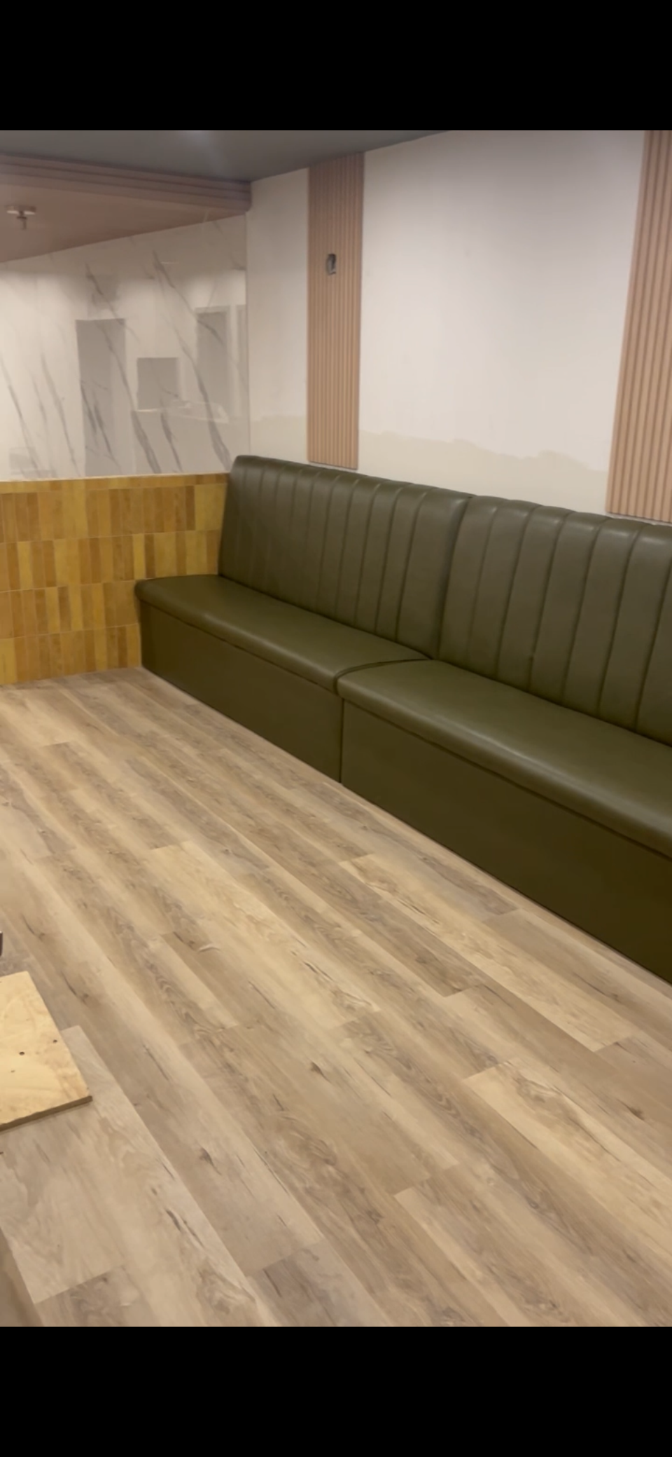 Interior of a room with a long green cushioned bench against the wall, wooden wall paneling, wood-look flooring, and a marble-like wall design in the background.