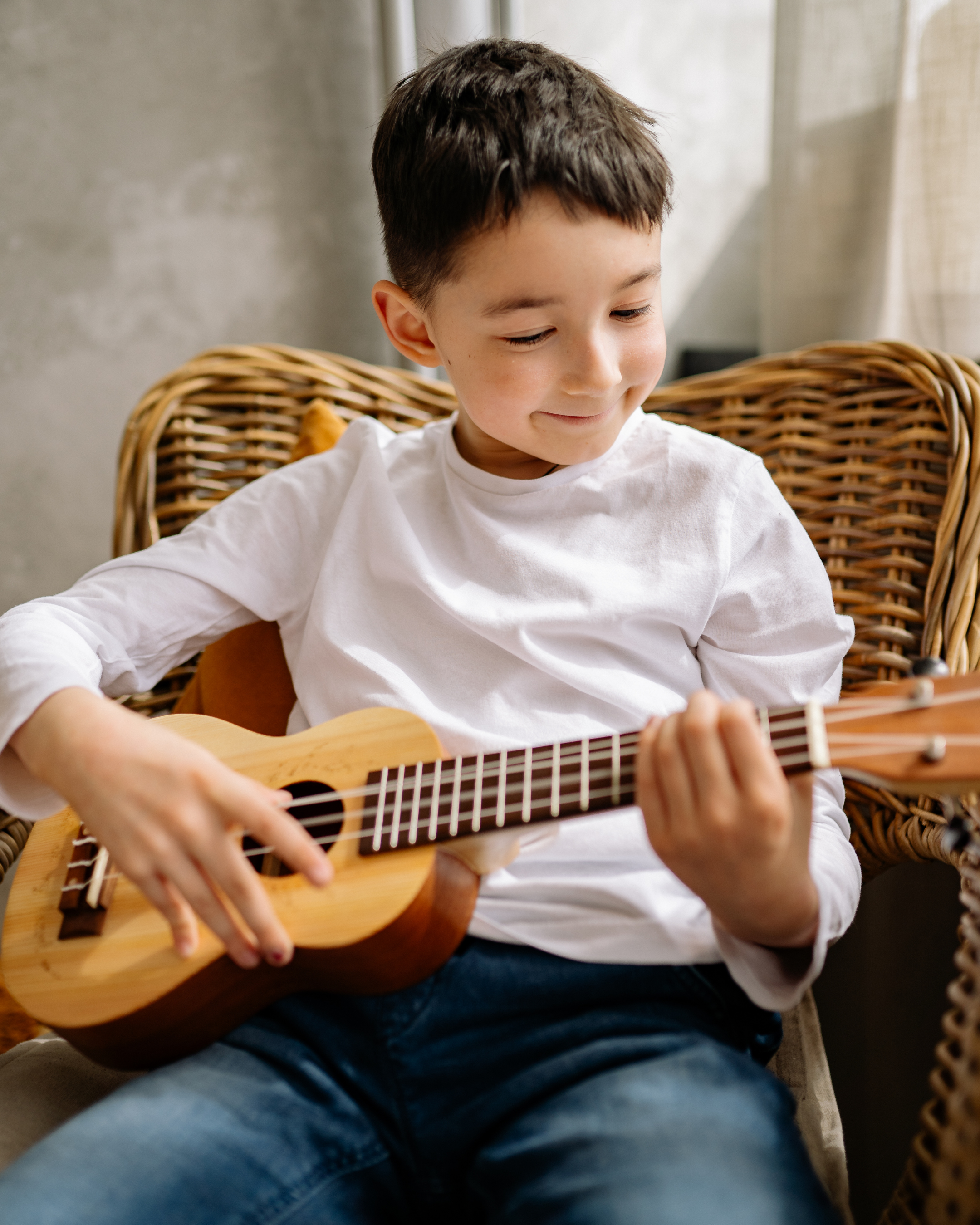 Young boy sitting in a wicker chair, playing a small wooden guitar and smiling.