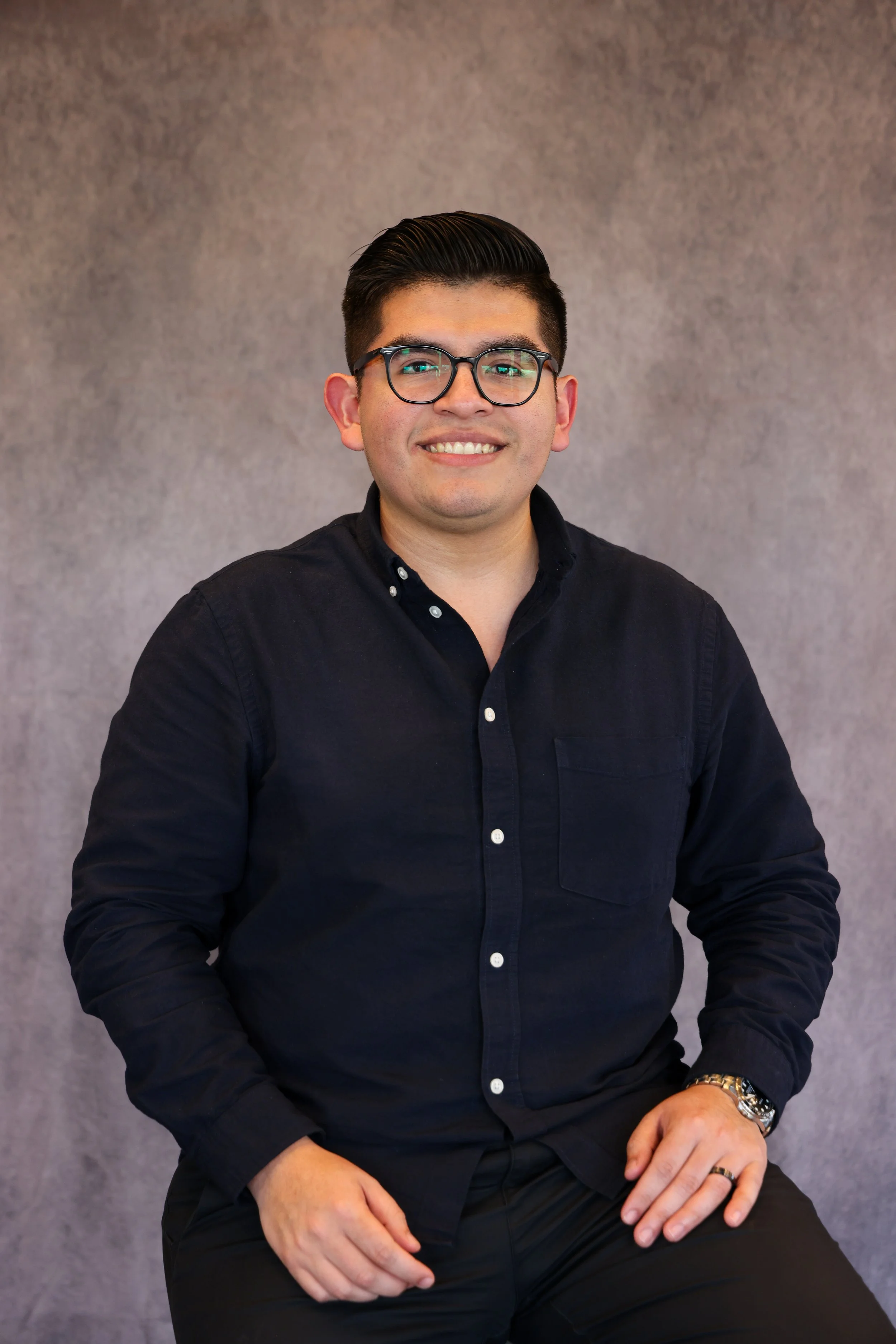 Portrait of a young man with dark hair, glasses, wearing a black button-up shirt, sitting against a neutral background, smiling at the camera.