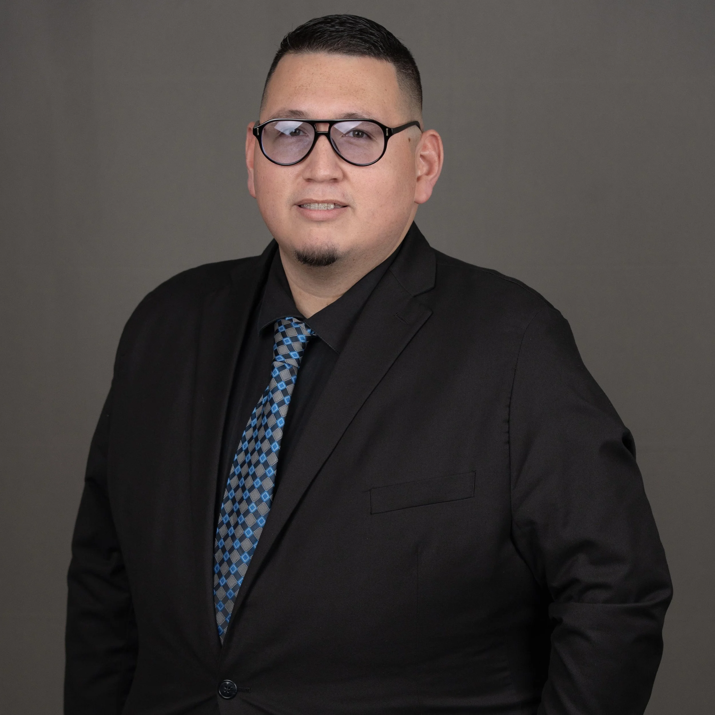 A man with glasses, a black suit, black shirt, and a patterned blue tie, posing in front of a gray background.