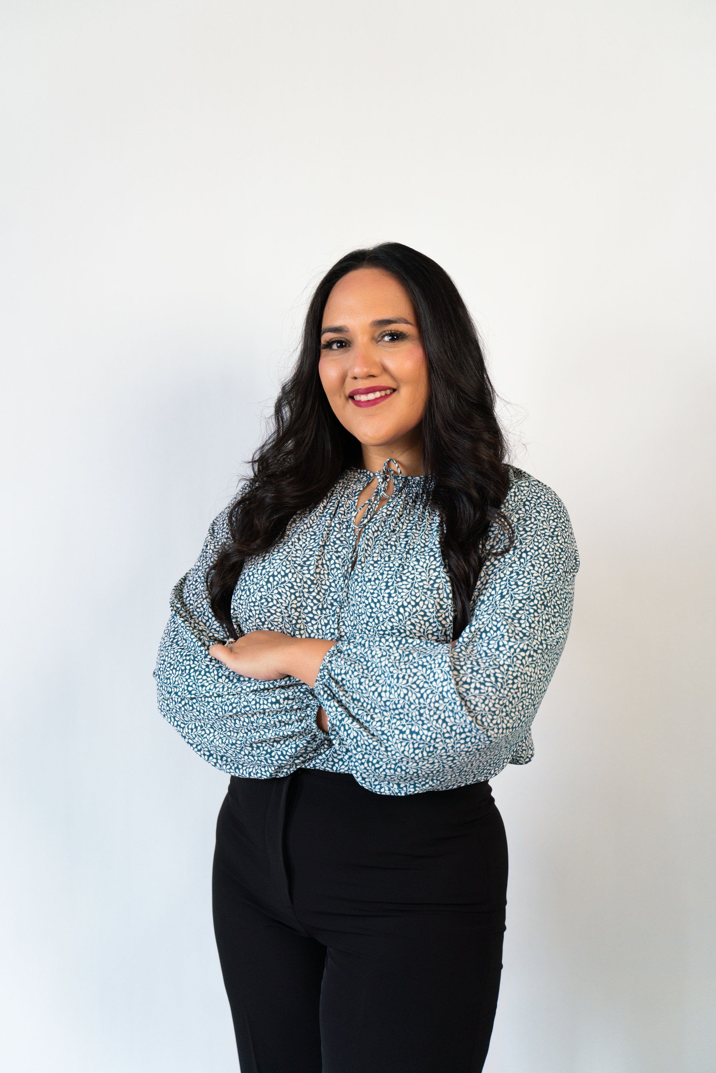 A woman with long dark hair, wearing a patterned blouse and black pants, standing with arms crossed, smiling in front of a plain white background.