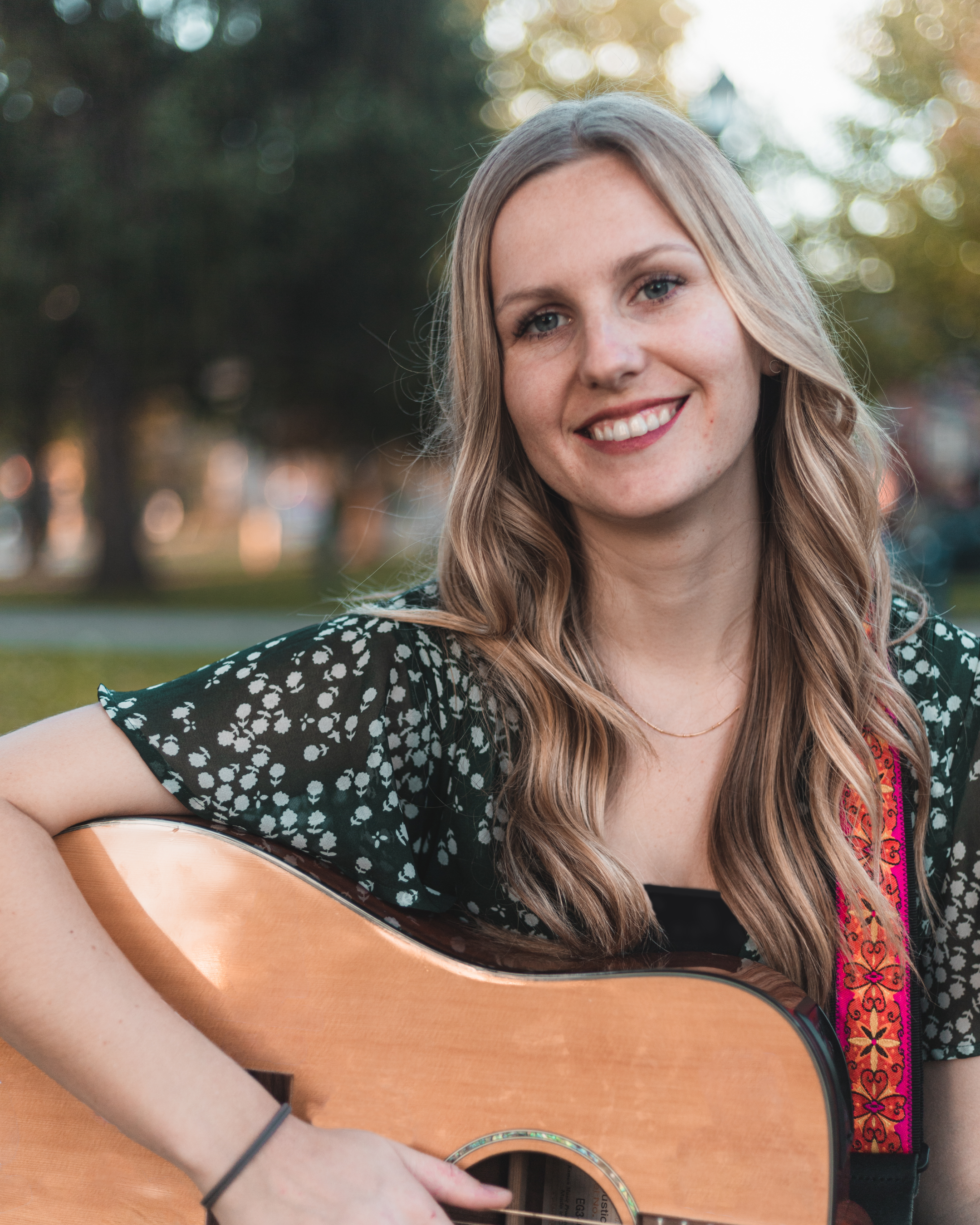 A young woman with long blond hair smiling and holding an acoustic guitar outdoors in a park during sunset.