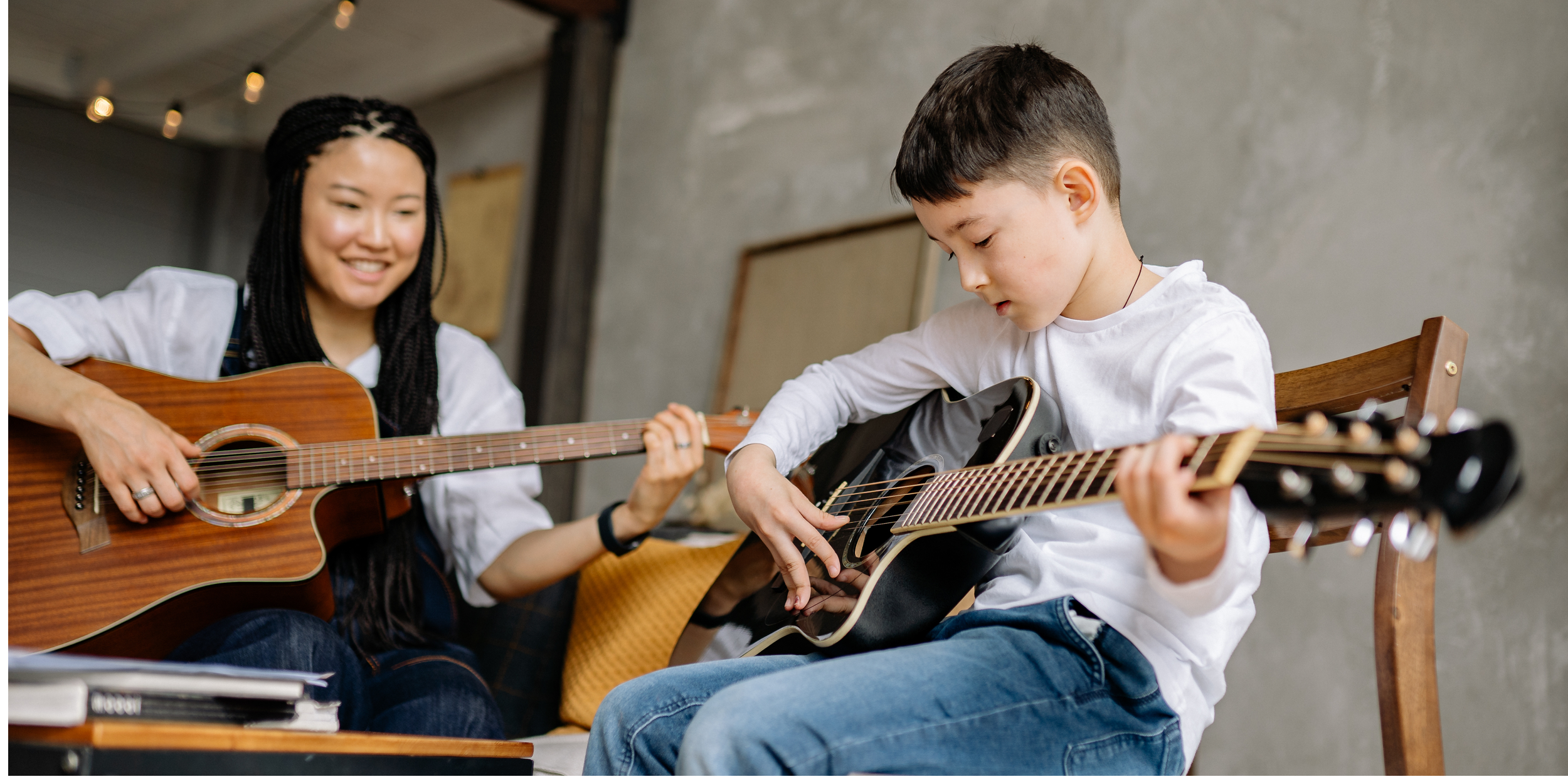 A smiling woman teaching a young boy how to play guitar. Both are holding acoustic guitars, and the boy is focused on playing his guitar.