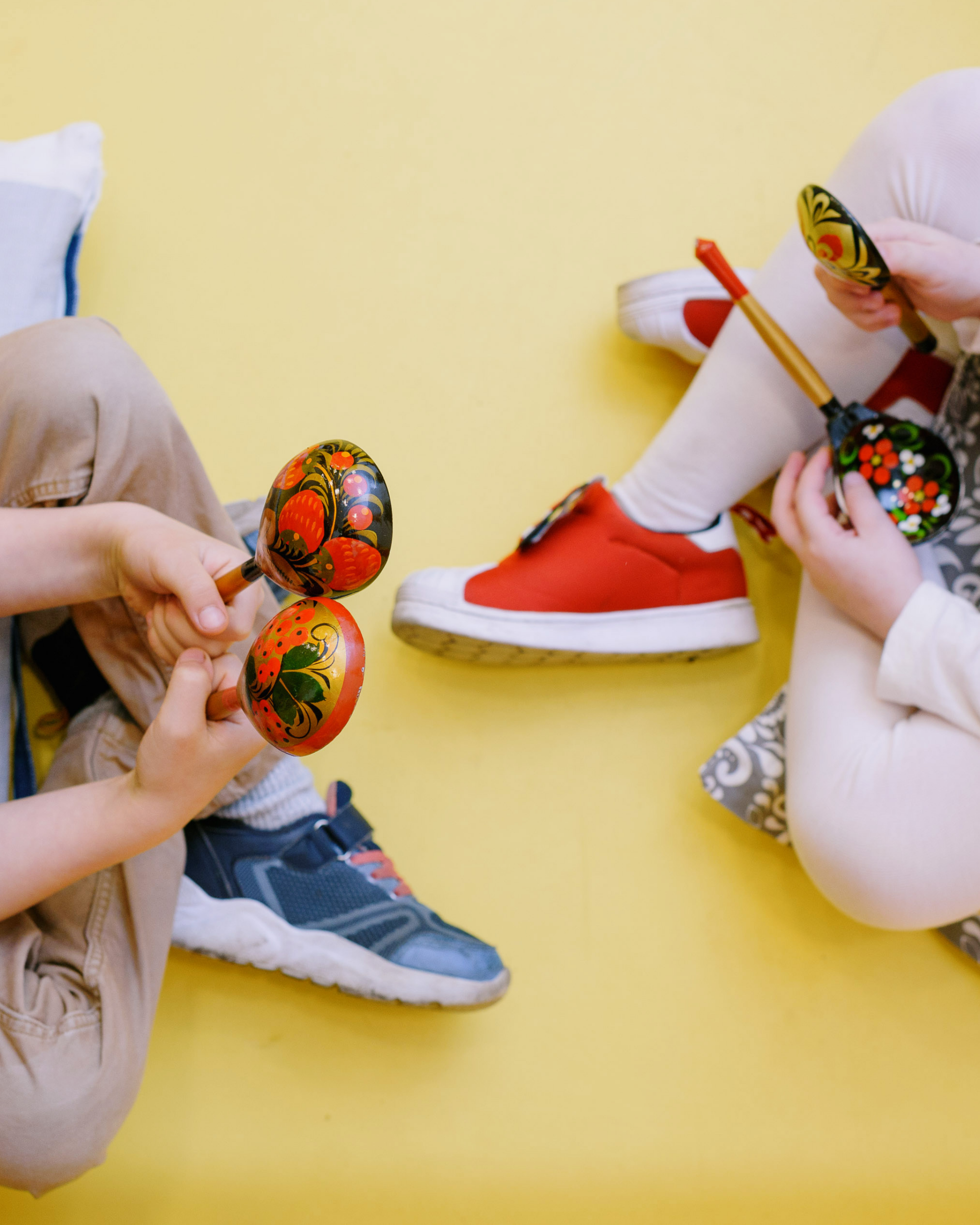 Two children sitting on a yellow floor, holding decorative painted instruments, with one child wearing red shoes and the other wearing sneakers.