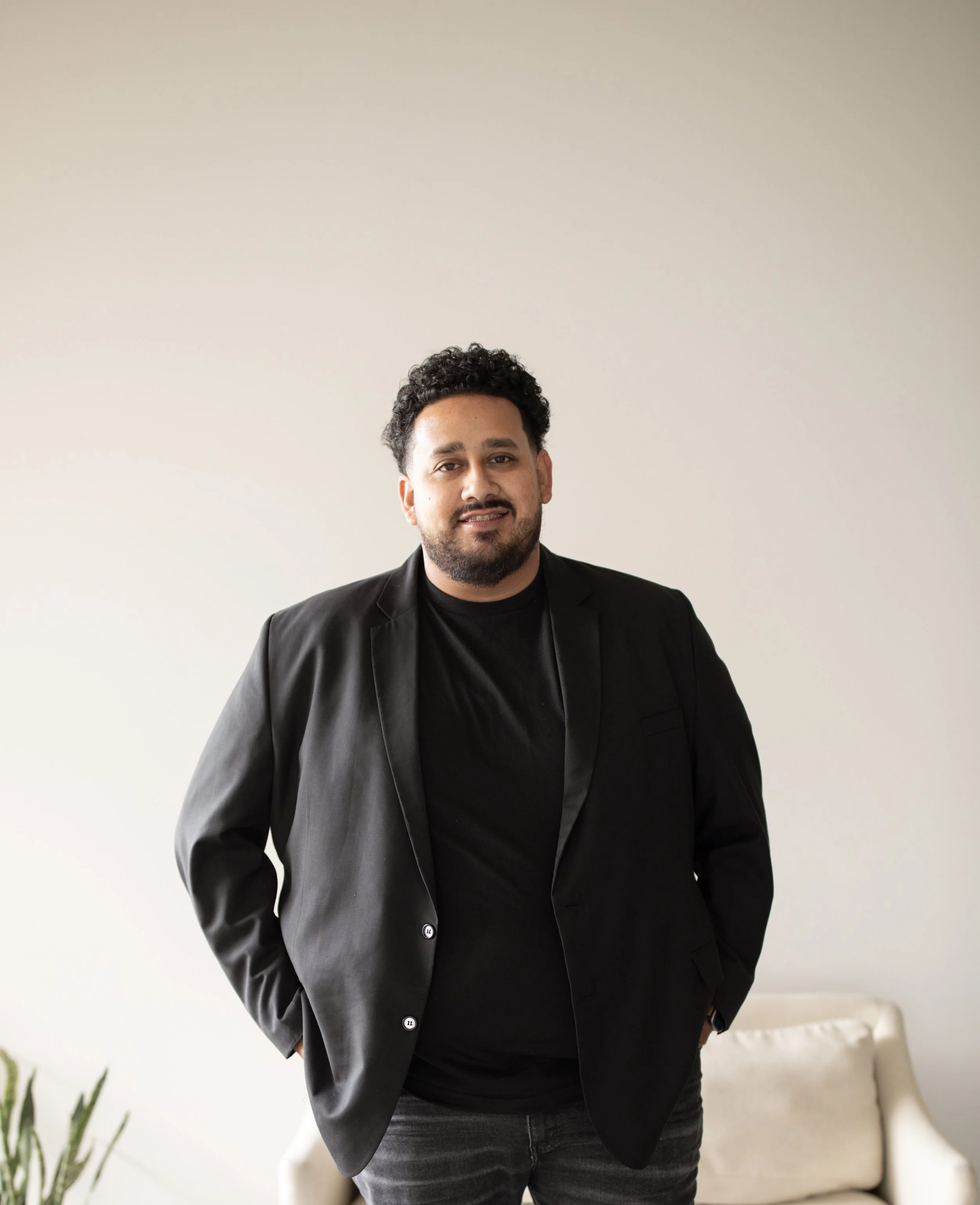 A man with dark curly hair and a beard wearing a black blazer and black t-shirt, standing in a room with a white wall and a cream-colored sofa behind him.