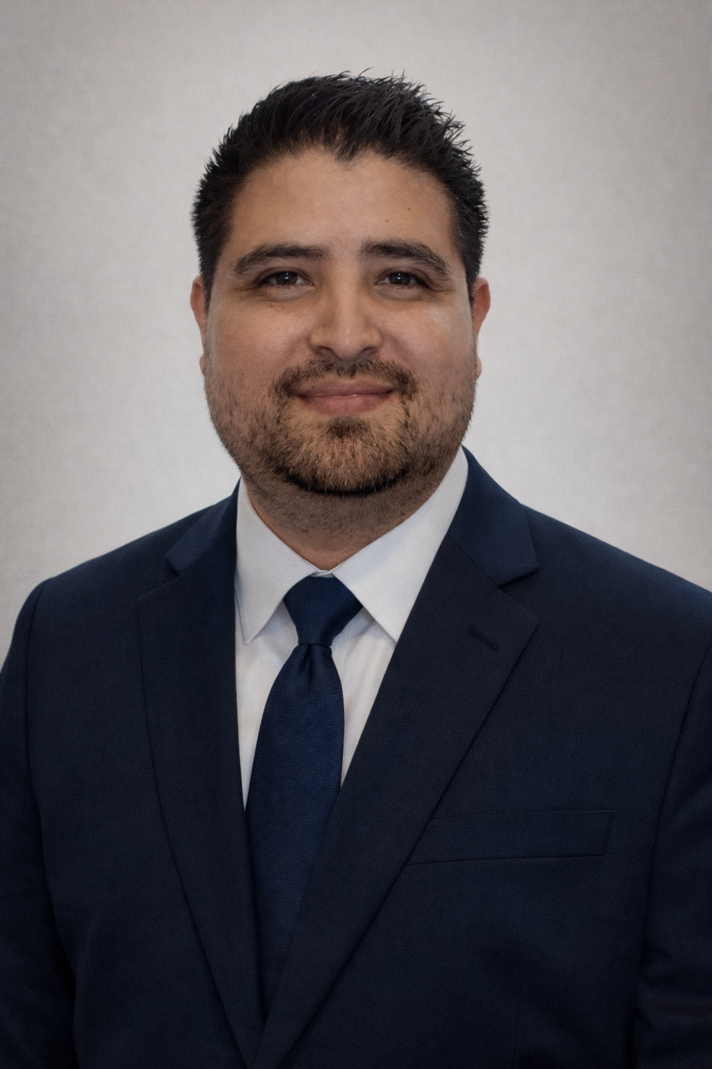 A professional man in a dark suit, white shirt, and dark tie, smiling with short dark hair and a beard, against a plain light background.