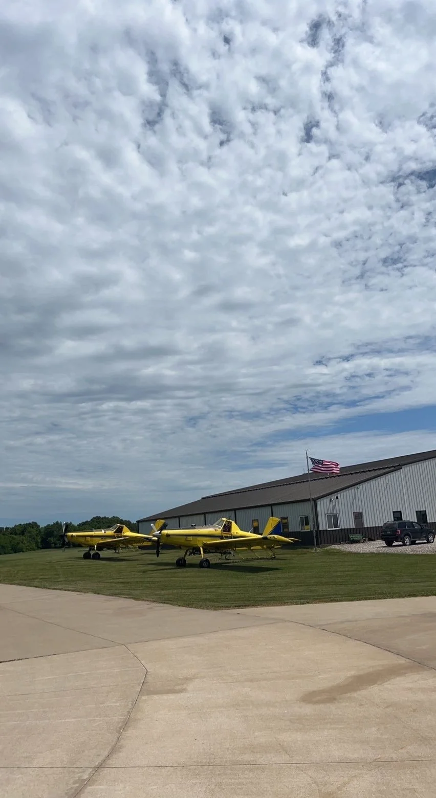 Two yellow training aircraft parked on the grass near a building with an American flag, under a partly cloudy sky.