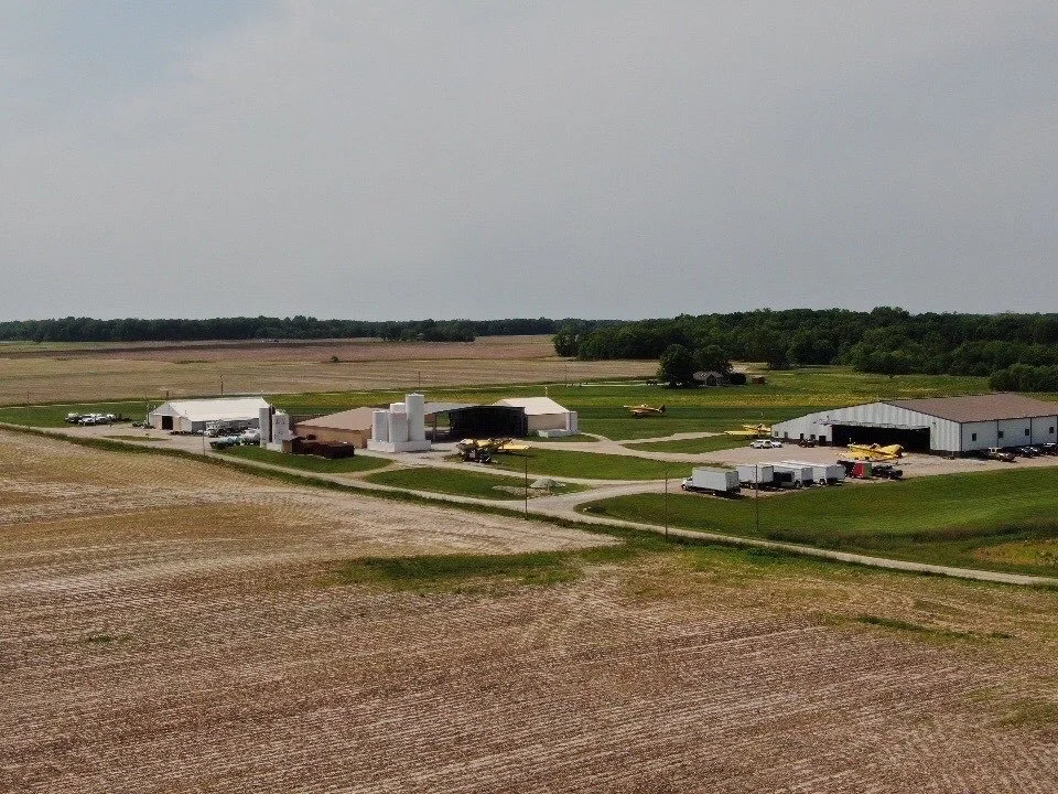 Small rural airport with a few small planes, hangars, and farm buildings surrounded by open farmland under a cloudy sky.