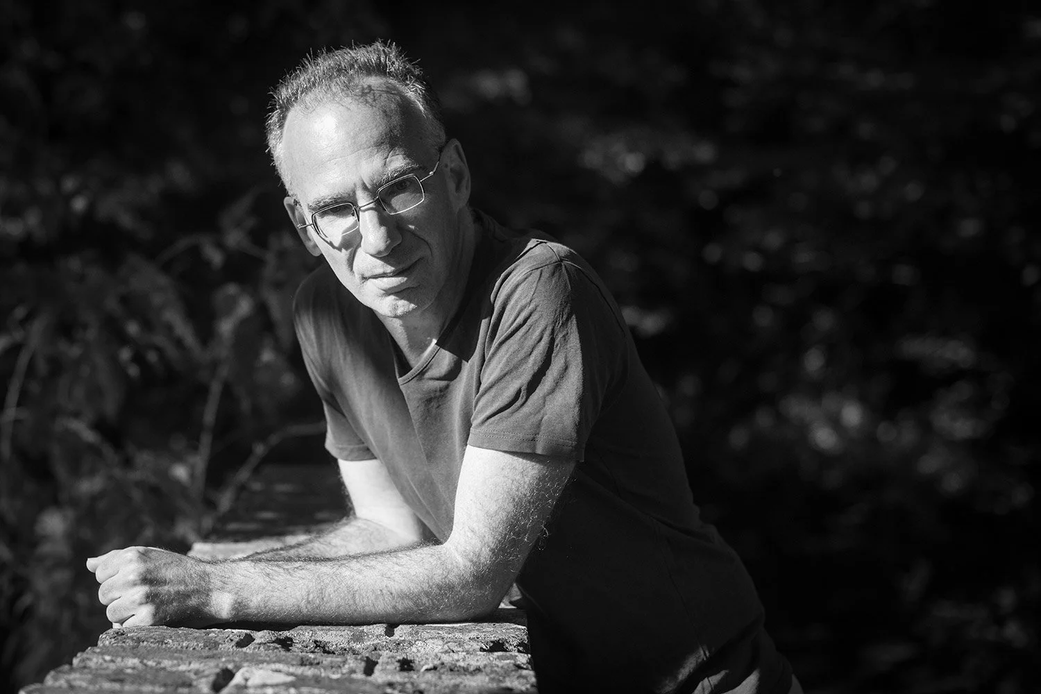 A black and white photo of a Yaron White, a BACP-registered bibliotherapist based in Stroud, Gloucestershire, leaning on a brick surface outdoors, with blurred foliage in the background.