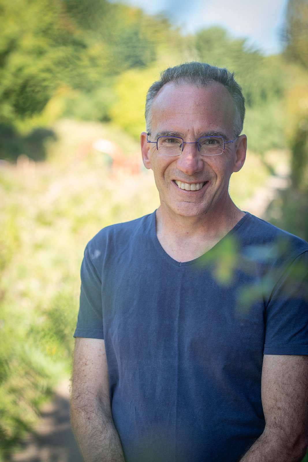 Yaron White smiling wearing glasses in a short-sleeved blue shirt outdoors, blurred greenery in the background near to Stroud Brewery.