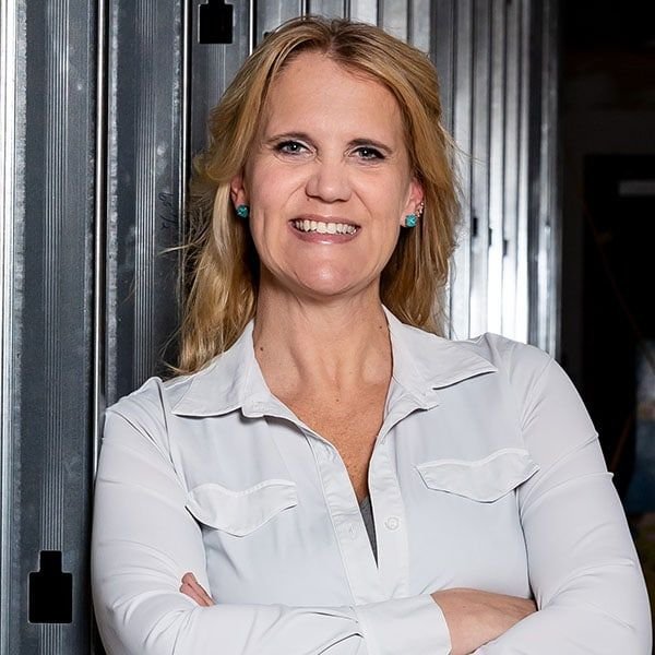 A woman with blonde hair wearing a white button-up shirt and turquoise earrings, standing with arms crossed in front of a metallic background.