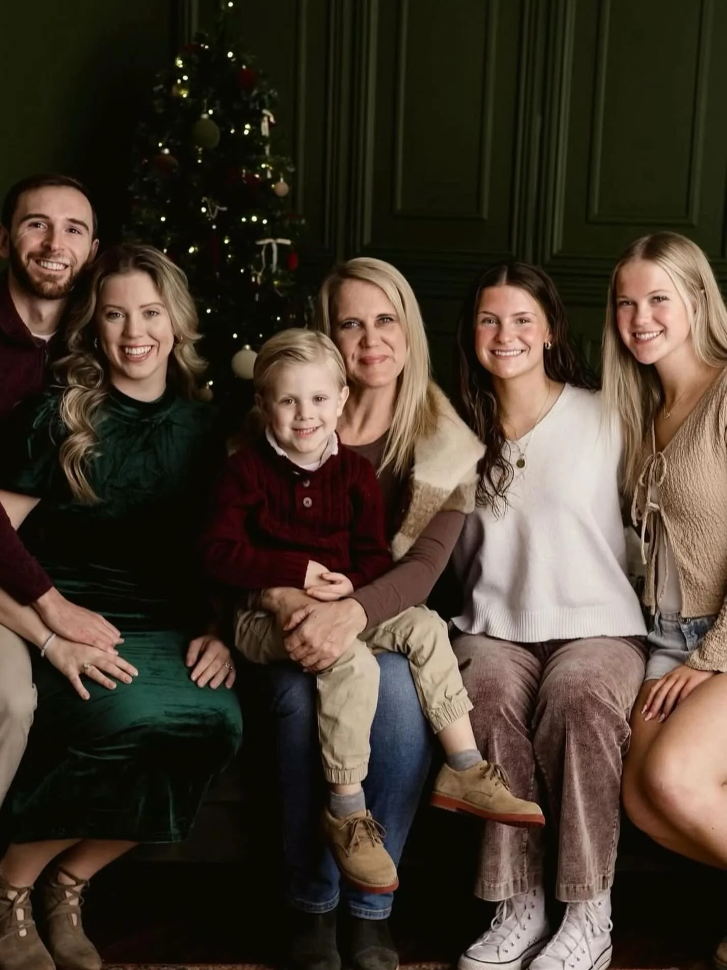 A family group photo during Christmas with a decorated Christmas tree in the background. The group includes six women, a man, and a young boy sitting in front. They are smiling and dressed casually, sitting close together in front of a dark green wall.