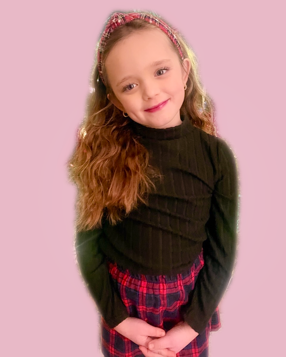 Delaney, a young girl with long curly hair, wearing a black ribbed turtleneck sweater, red plaid pants, and a matching headband, smiling softly against a pink background.