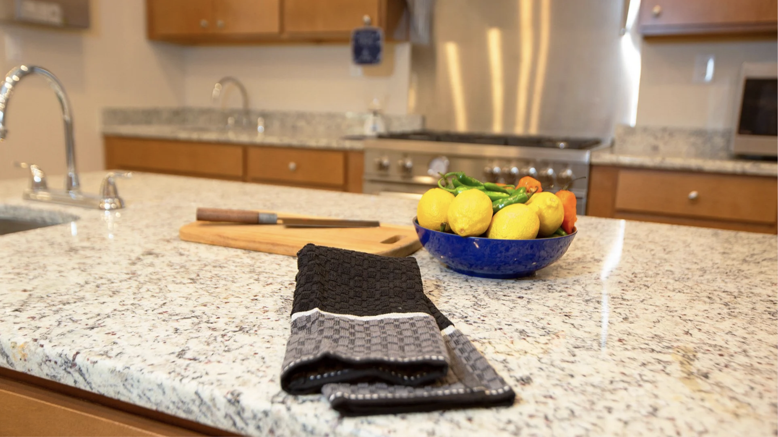 Kitchen countertop with a bowl of lemons and peppers, a wooden cutting board, a black kitchen towel, and a knife, with a stove and cabinets in the background.