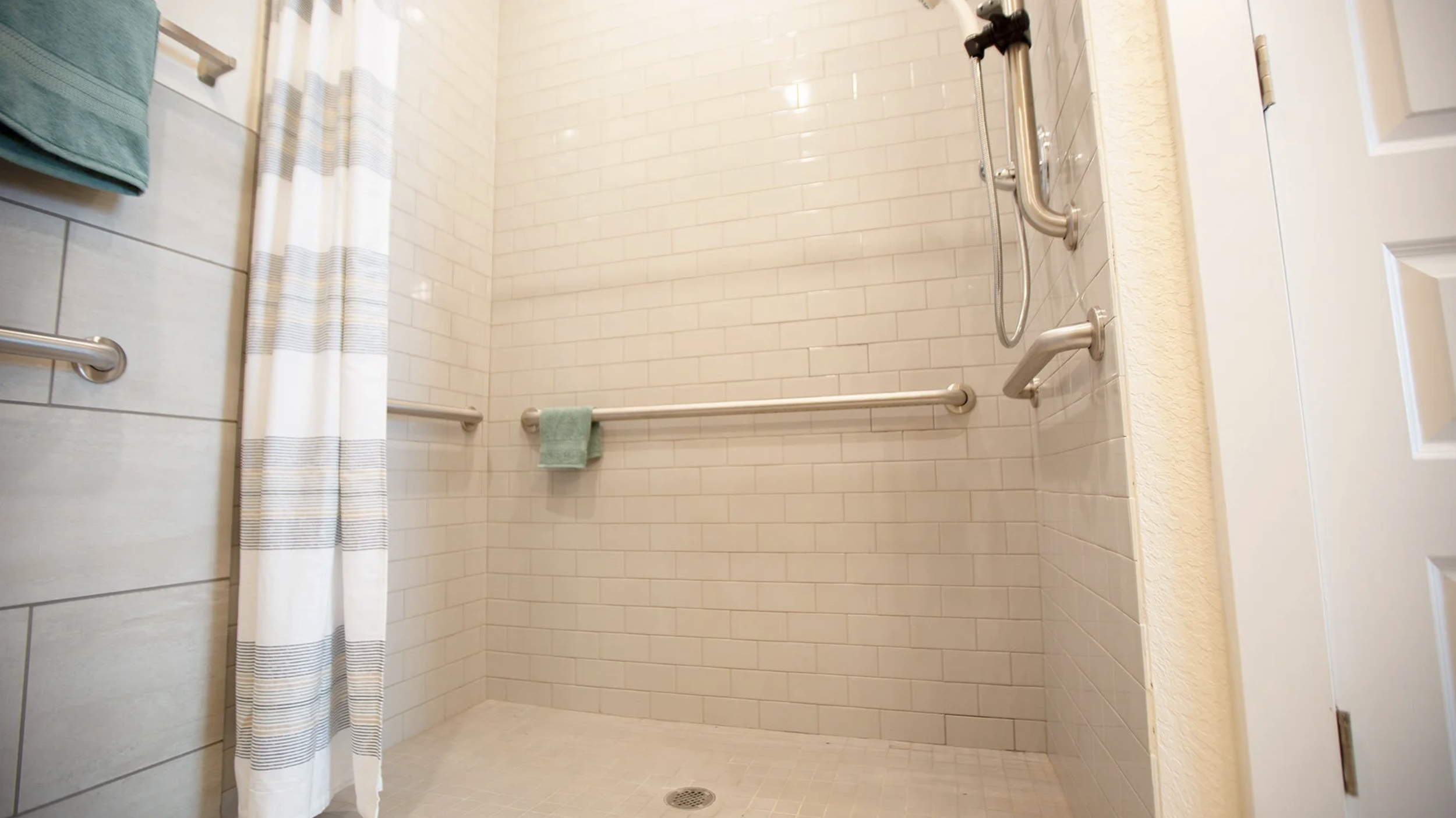 Accessible walk-in shower with white tile walls and beige floor, equipped with grab bars, a fold-down seat, and a handheld showerhead.