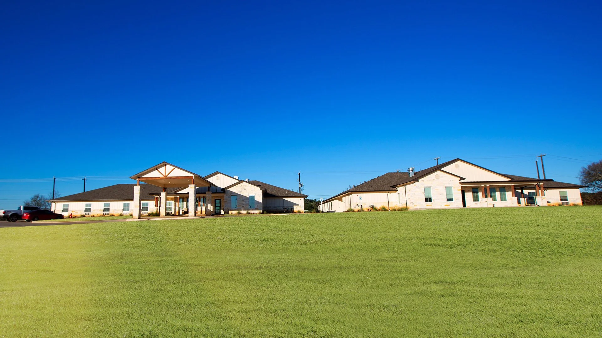 Modern single-story building with a stone exterior, large windows, and a covered entrance, set against a clear blue sky and green grass.